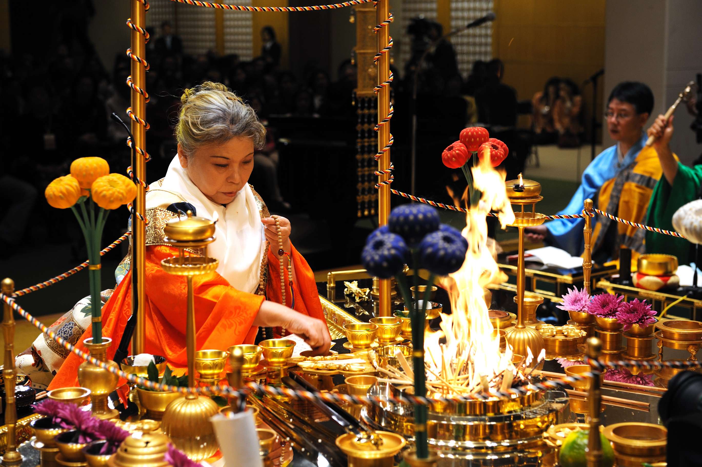 Her Holiness in orange ceremonial robes sits at a fire ceremony altar filled with golden ritual articles, offerings, and surrounded by a colored cord, reaching for something near a large flame.