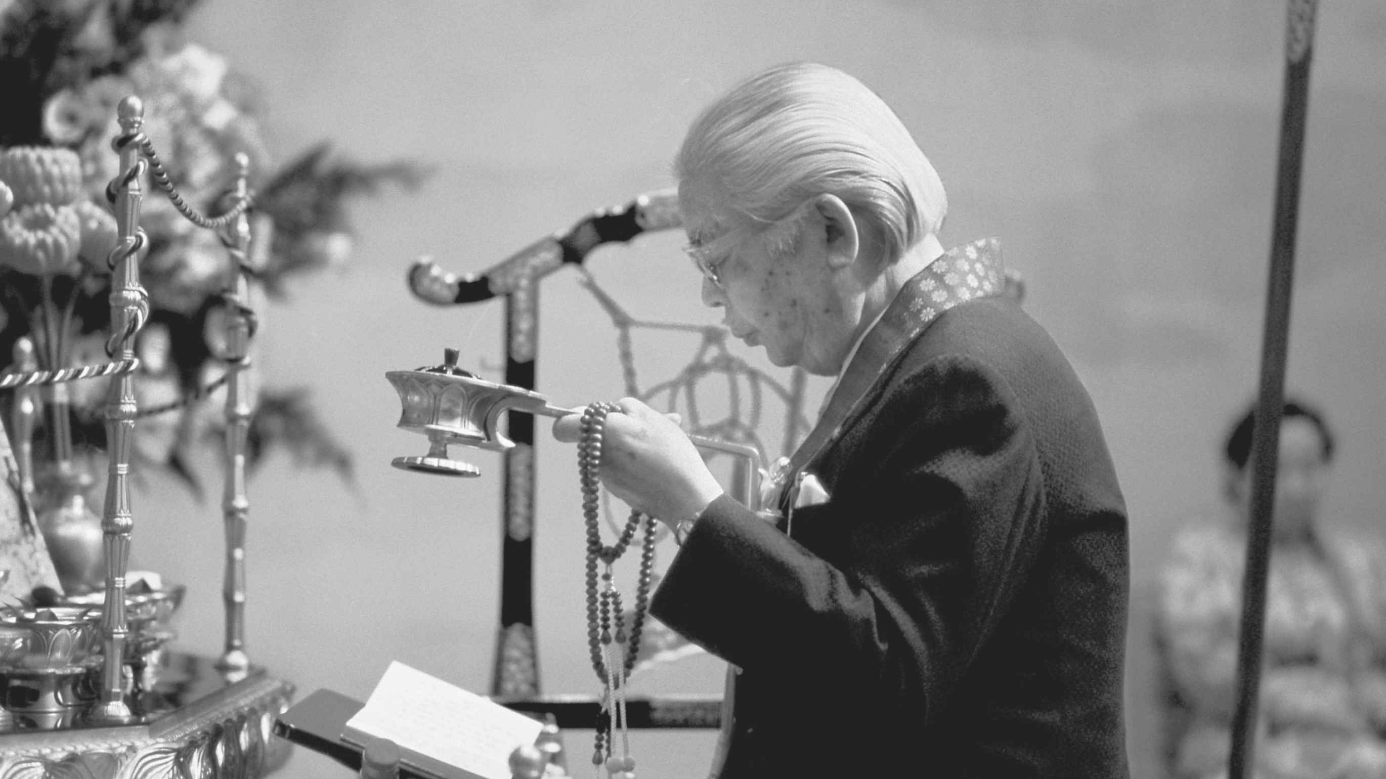 A white-haired Japanese man wearing a dark suit jacket with a brocade priestly surplice around his neck and holding a censer and rosary in his left hand reads from a book of prayers in front of an altar laden with ornate symbolic offerings.