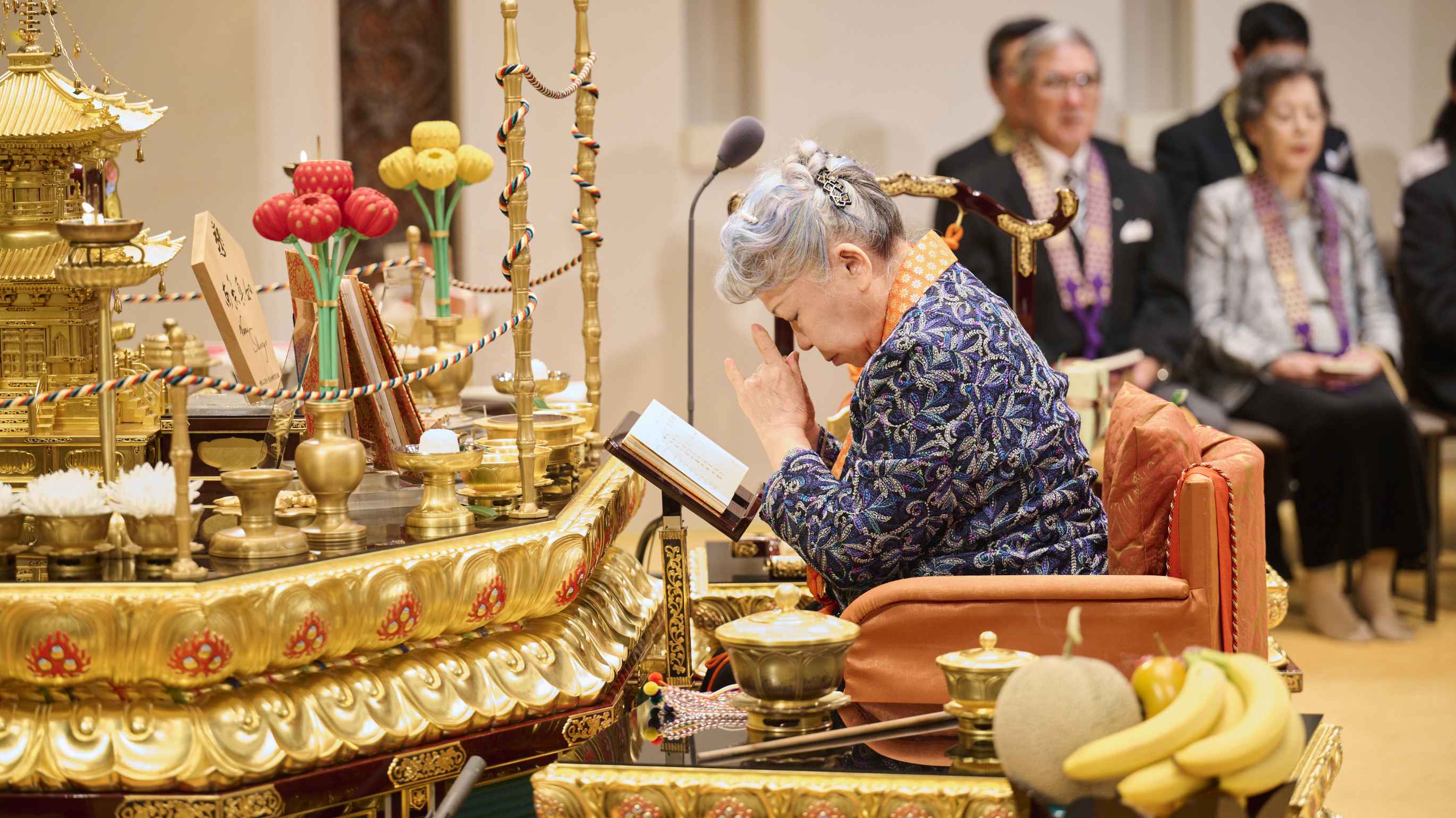 A gray-haired Japanese woman wearing an orange, brocade priestly surplice around her neck, sits in a chair before an ornate altar laden with symbolic offerings, bowing her head and forming a mudra with her hands by pressing her little- and fore-fingers together with the others folded in as she prays; others rapt in prayer are seated in the background.