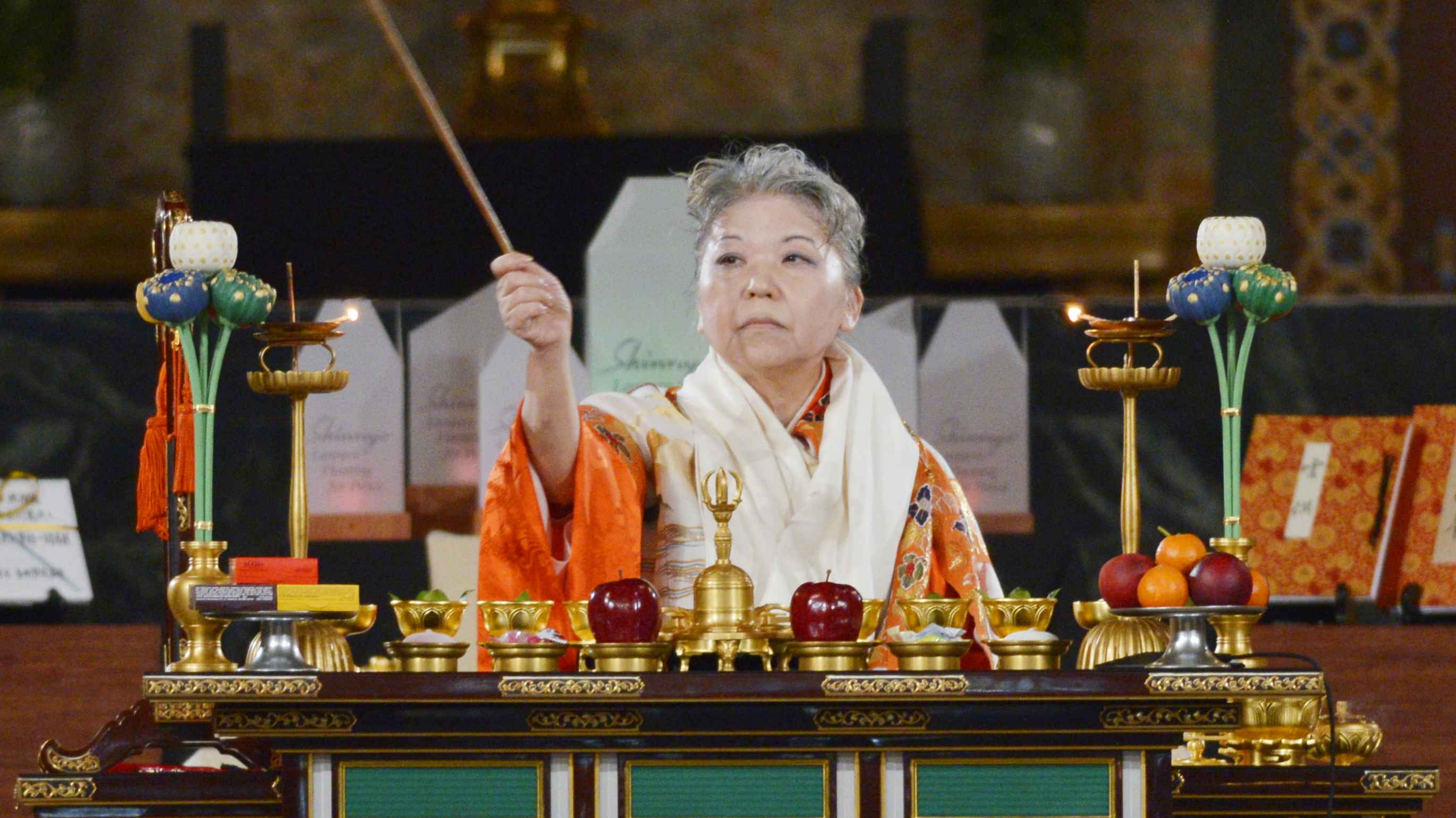 A gray-haired Japanese woman seated before a table bearing ornate symbolic offering bowls, arranged fruit, and ritual implements, wearing vermillion priestly robes and a white scarf around her neck, raises her right arm to wield a long wand-like ritual implement as she performs a blessing rite.