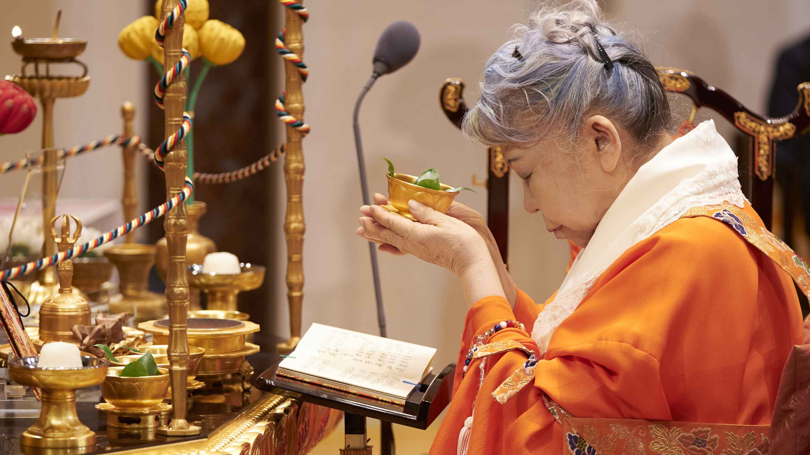 A gray-haired Japanese woman with her eyes closed in prayer and wearing vermillion priestly robes with a lacy white collar is seated before an ornate altar laden with symbolic offerings, as she lifts a small golden bowl holding green leaves in a gesture of offering.