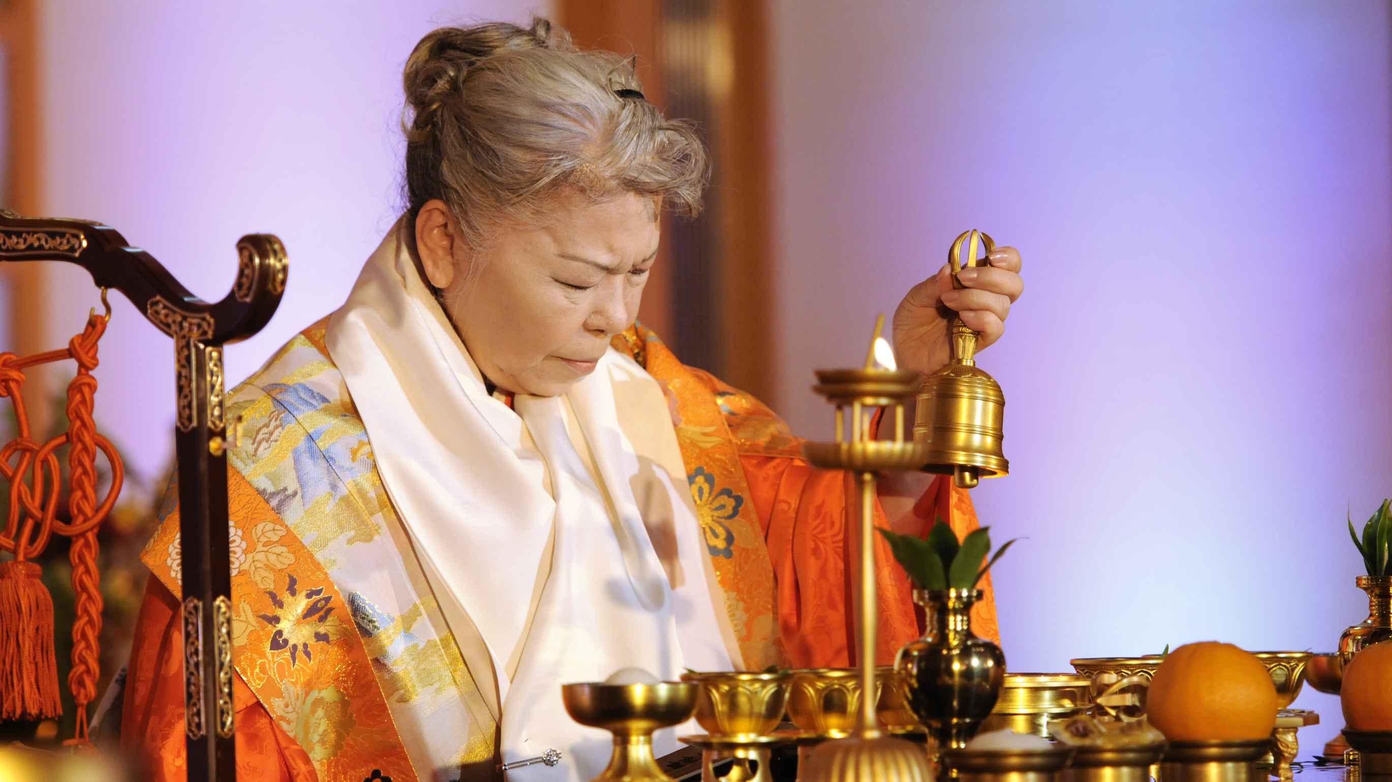 A gray-haired Japanese woman clad in vermillion priestly robes with a white silken scarf around her neck sits with her eyes closed as if in concentrated prayer as she wields a golden hand-held bell in her left hand before a table on which a series of golden offering bowls, some holding fruit, others holding leaves, are arranged.