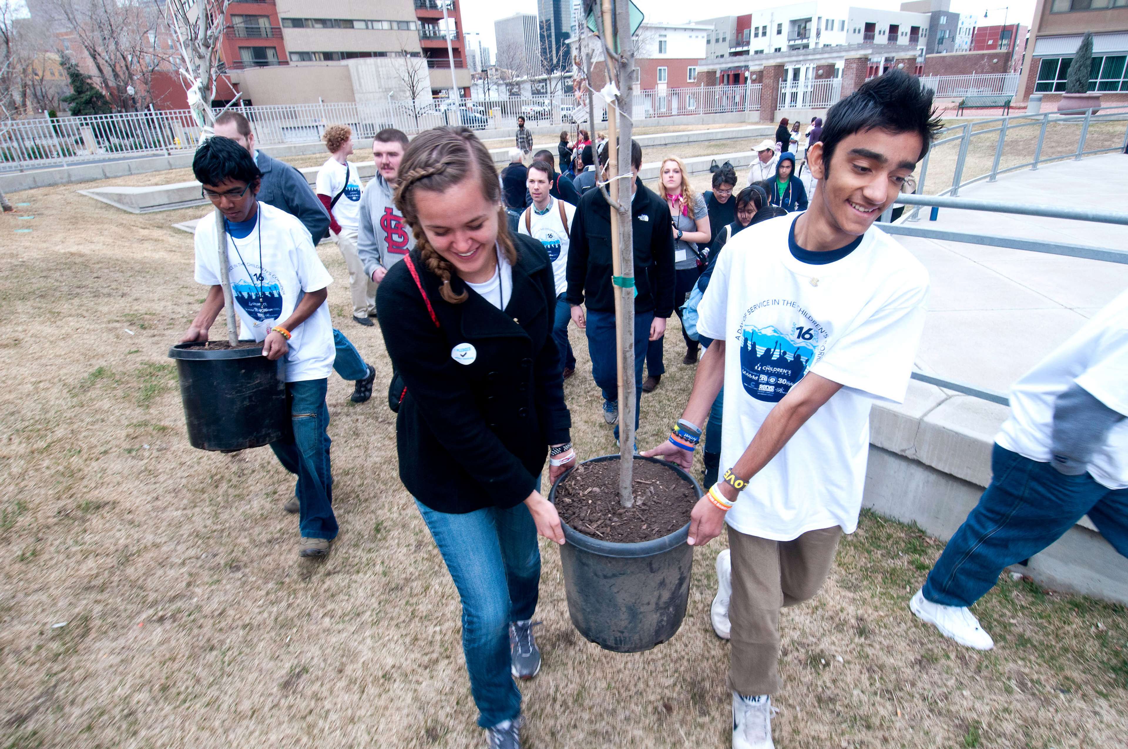 A young man and woman work together to carry a sapling in a bucket in an outdoor, urban greenspace; other young people carrying saplings follow behind.