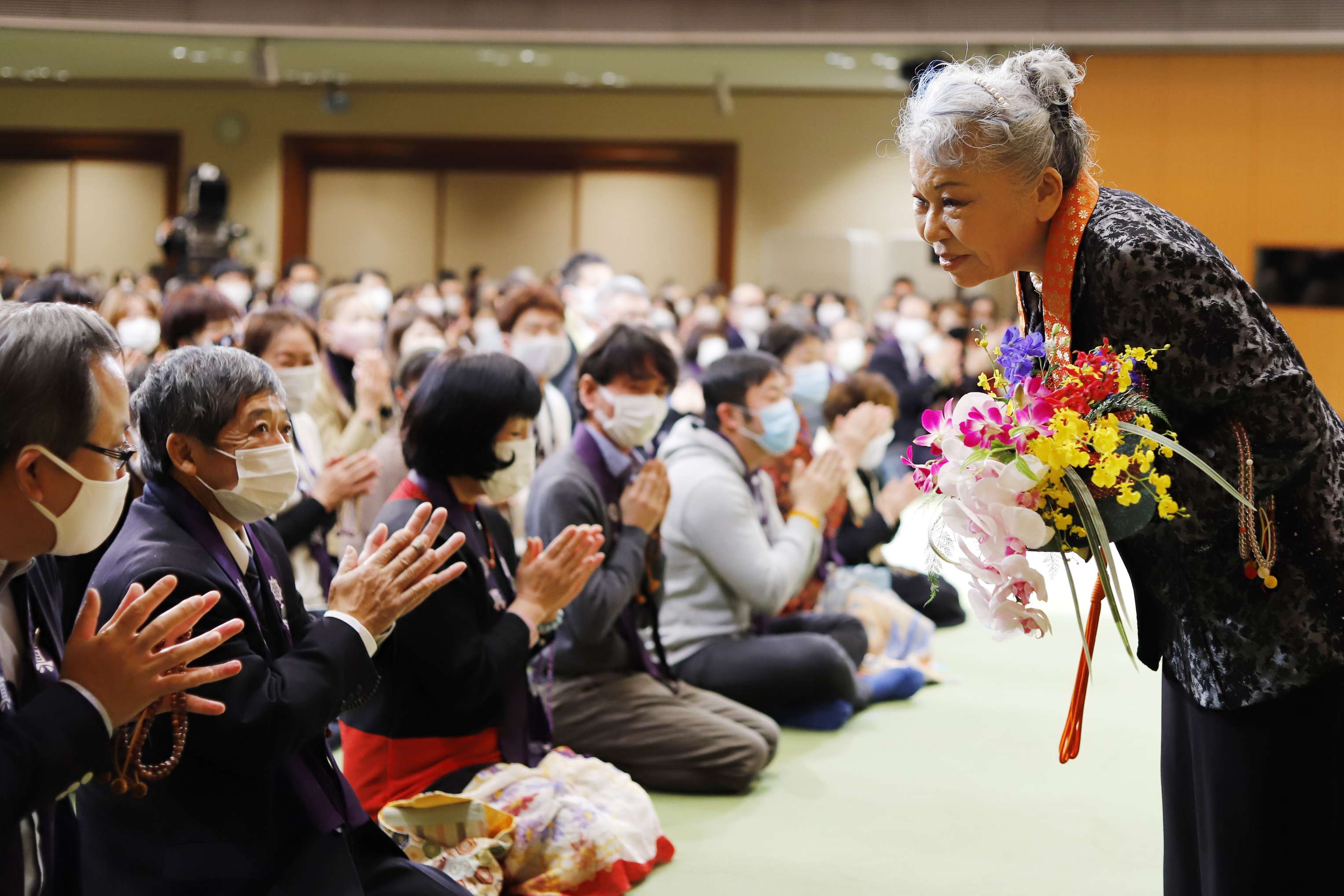 Her Holiness and a diverse group of people, all smiling, laughing, and standing in a circle, place their hands together one on top of the other.