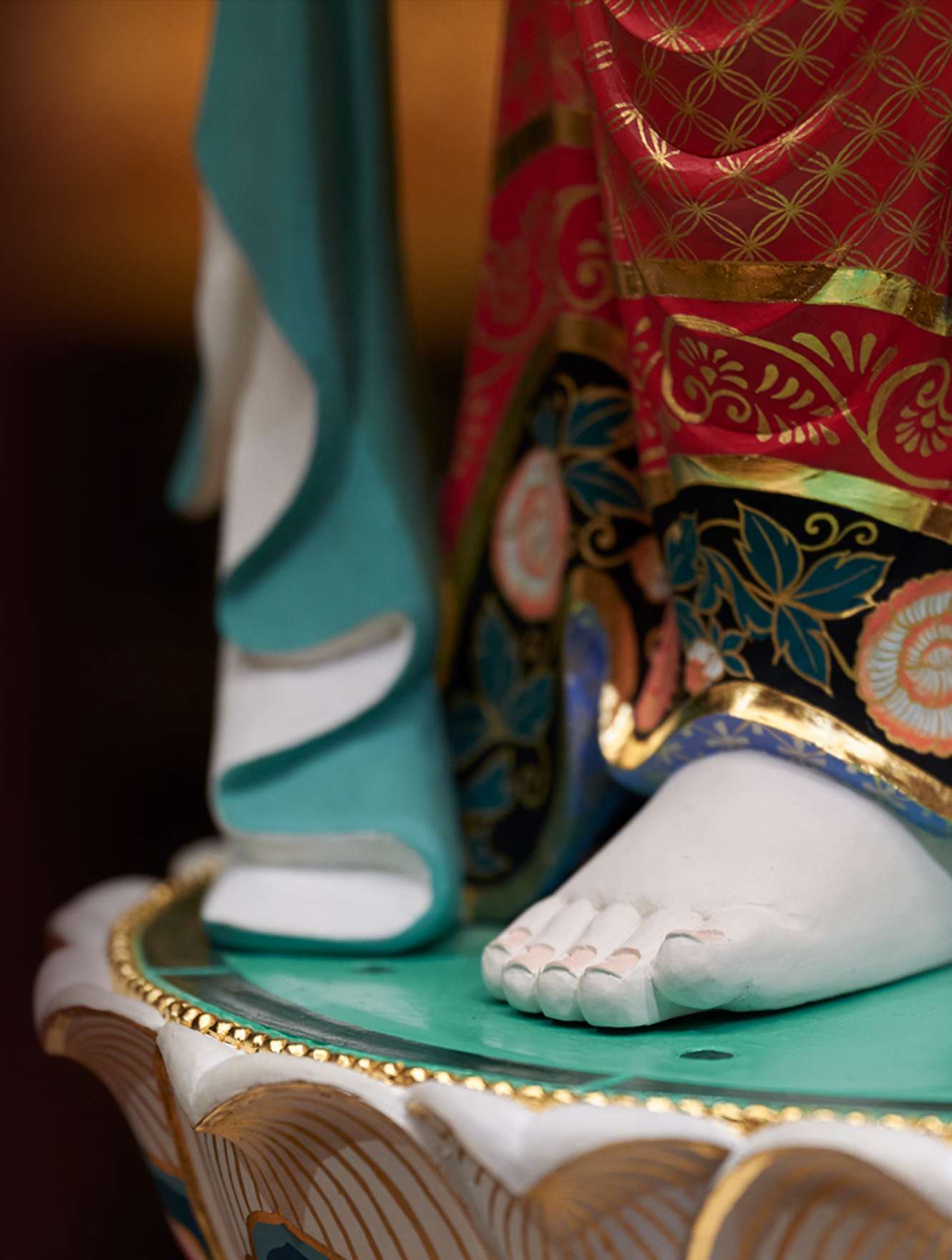 A cropped image of a statue shows a flowing garment of red and blue, intricately decorated with gold floral patterns, hanging above a soft, white porcelain foot that stands atop a lotus-shaped pedestal.