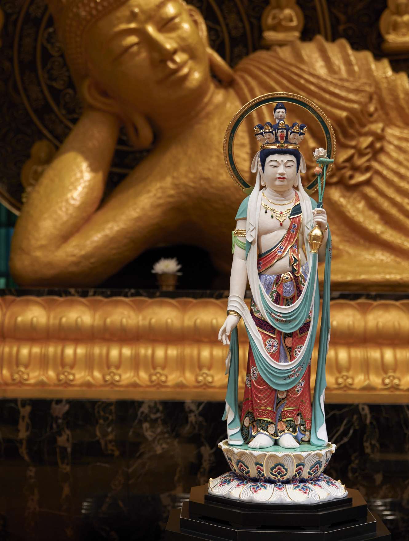 A full length statue of a pale, serene, standing figure draped in colorful silken garments, adorned with a crown displaying ten smaller heads and aureole, holding a vase with lotus bud in its left hand, with its right hand hanging gracefully at its side is displayed before a large golden statue of the Buddha reclining at the moment of nirvana, a portion of which is visible in the background.