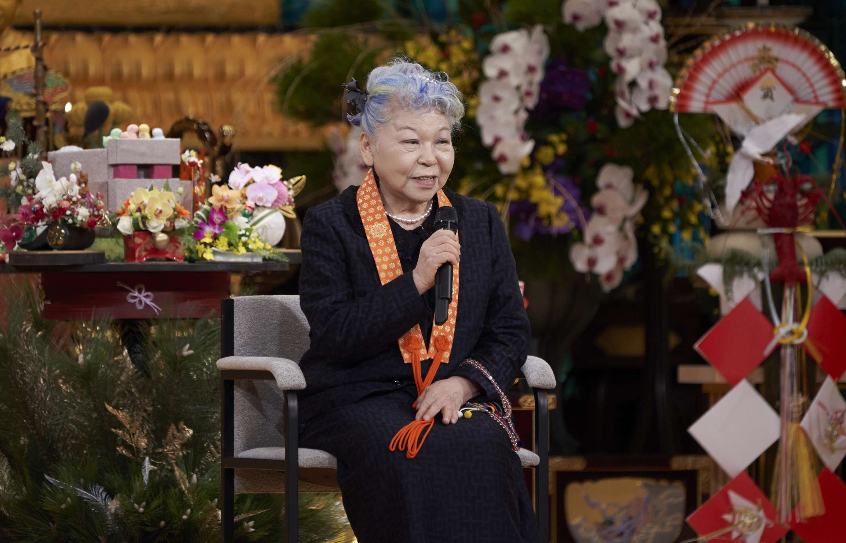 A gray haired Japanese woman wearing a dark suit jacket and dress, sits casually, facing the camera in front of an altar laden with flowers and other offerings, speaking into a microphone held in her right hand.