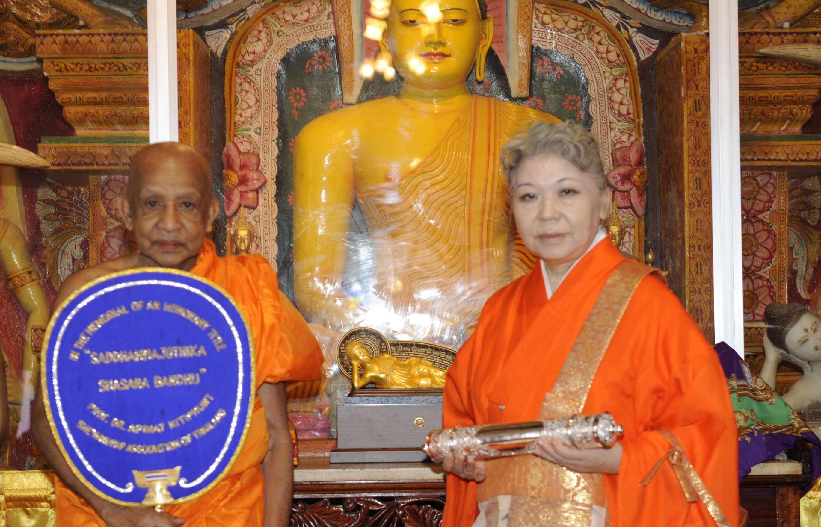 Her Holiness, in an orange robe, holds a golden cylinder, standing next to a shaven-headed monk who holds a large ceremonial fan decorated with words, in front of a large statue of Buddha.