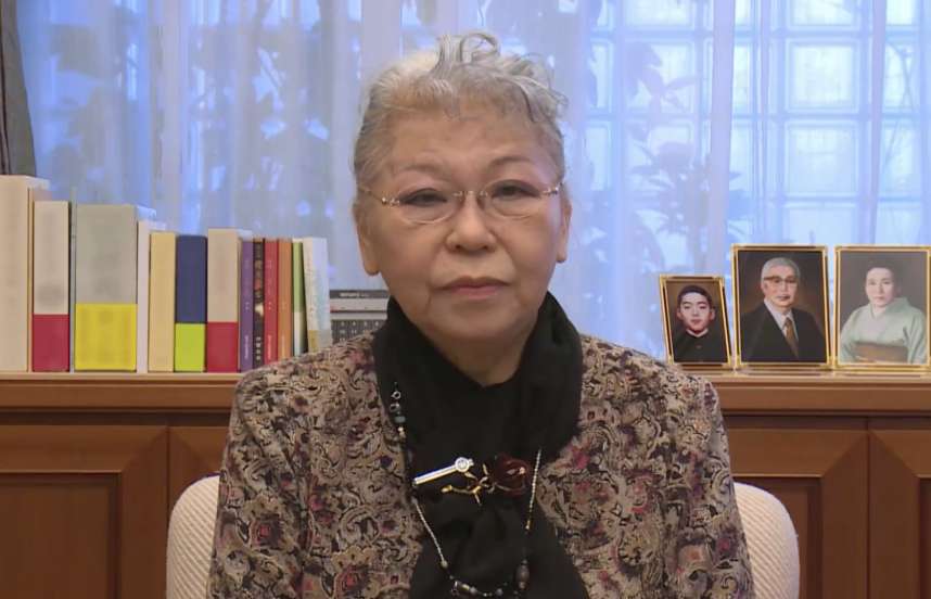 Her Holiness Shinso Ito, wearing spectacles, a patterned blouse with a black scarf, sitting in front of a window with a shelf displaying photos, looks directly into the camera.