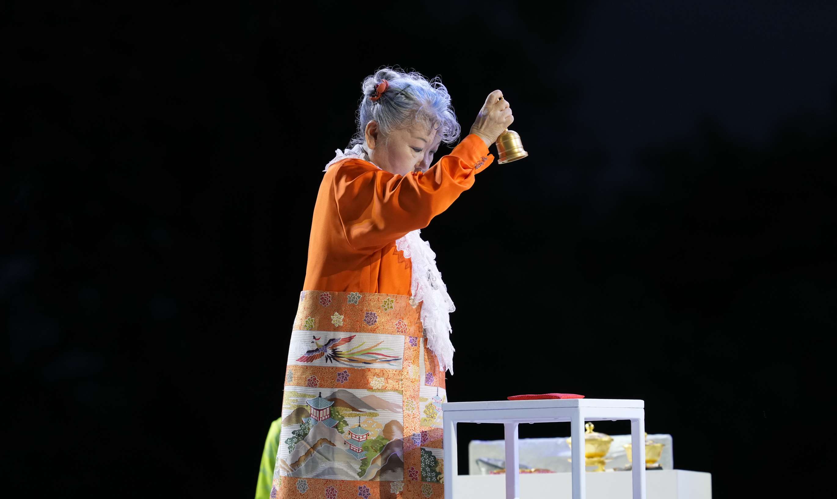 A elder Japanese woman, standing before a small low table holding implements, dressed in orange robes with an ornate brocade apron decorated with images of birds, solemnly holds a golden bell aloft in her outstretched right hand as she rings it; her face looks downward, as if she is inwardly in contemplation.