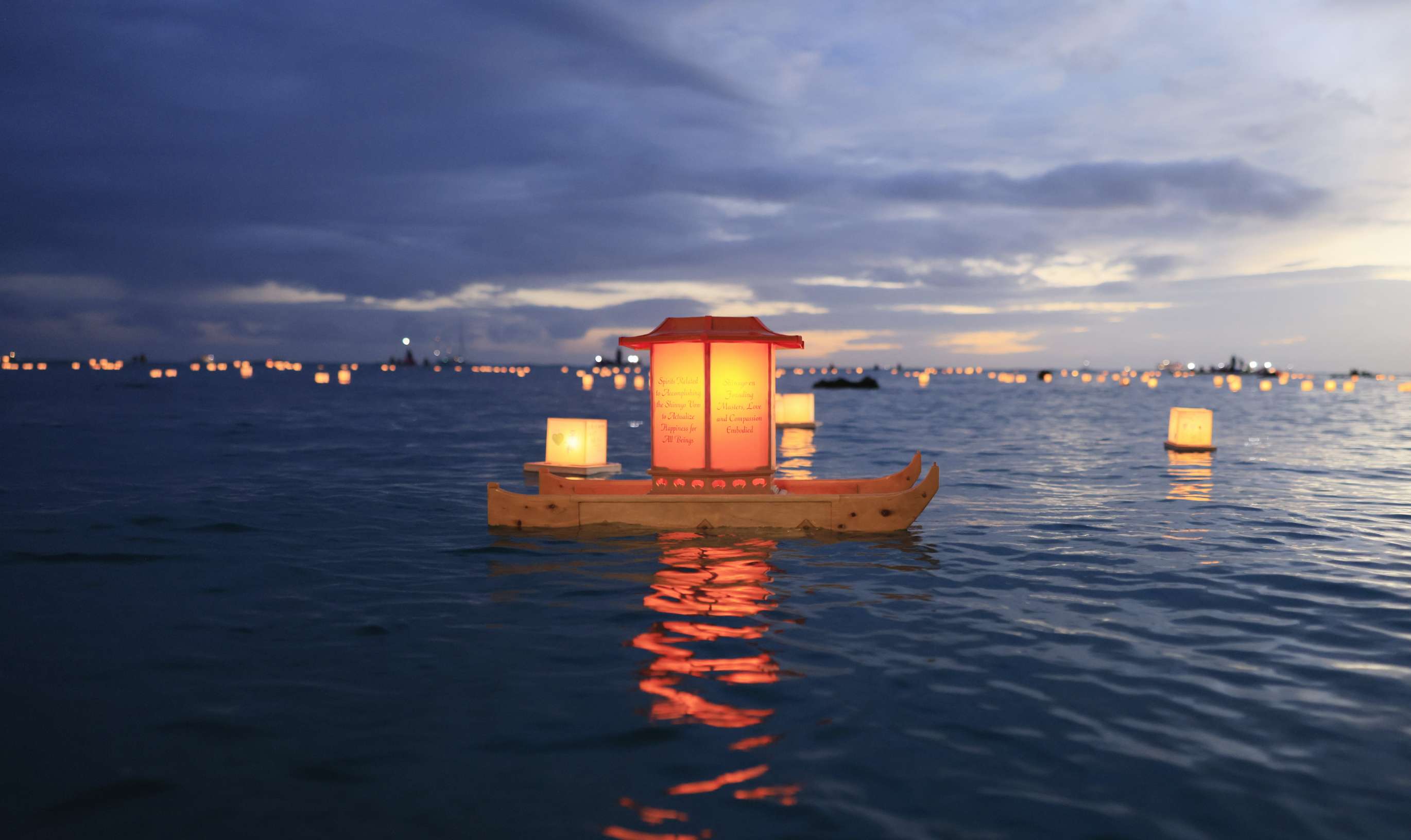 A large, traditional Japanese floating lantern sits atop the calm ocean waters of a bay at dusk; the sky darkens in the background and the orange glow of the lantern is reflected in the ripples of the water.