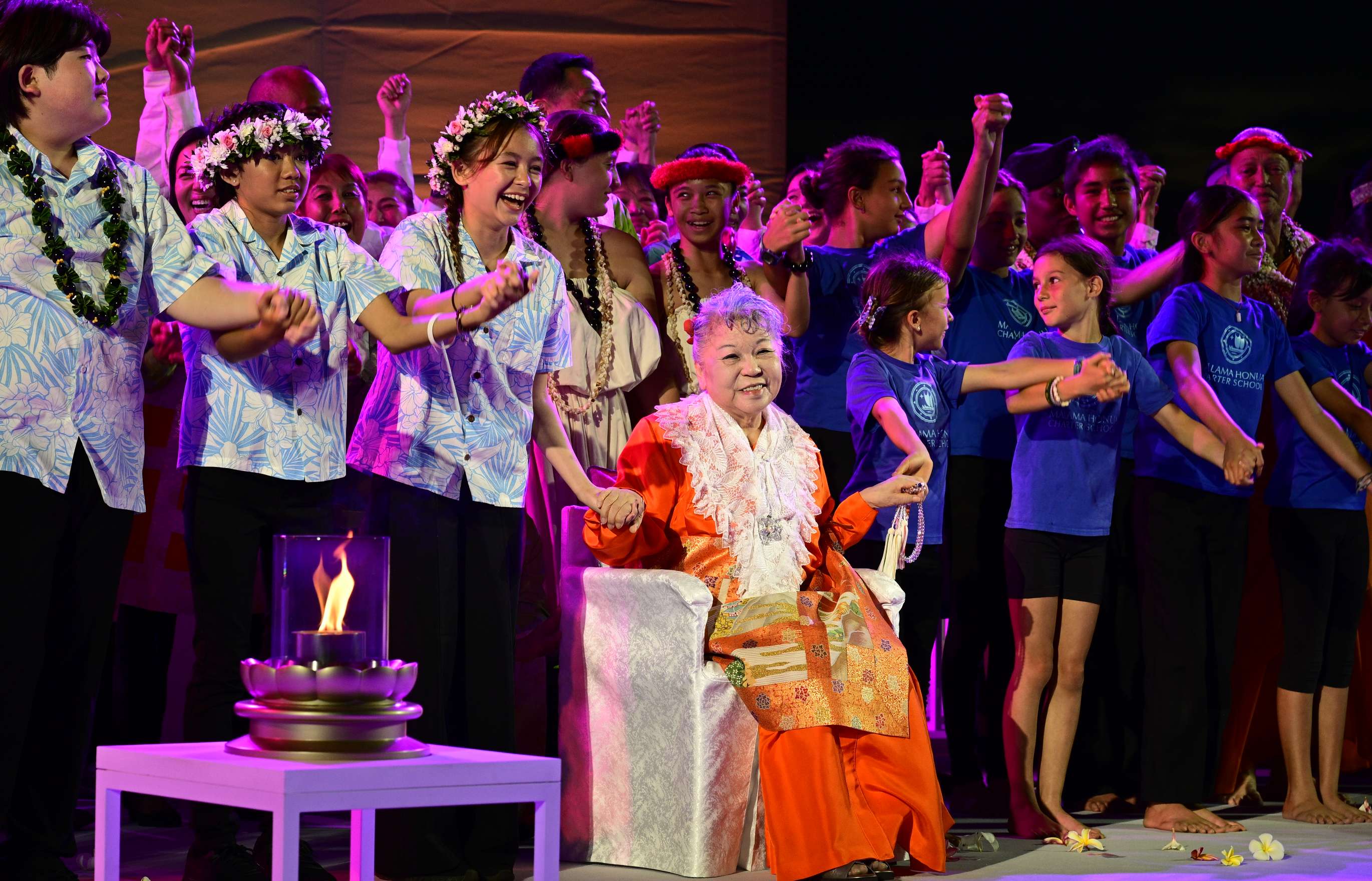 A smiling, elder Japanese woman, dressed in orange robes with a brocade apron and white shawl, sits on a chair under lights on a stage, holding hands with children and young-adults, who are all holding hands and joyfully smiling; a single open-flame lamp sits atop a lotus shaped base on a table in the foreground.
