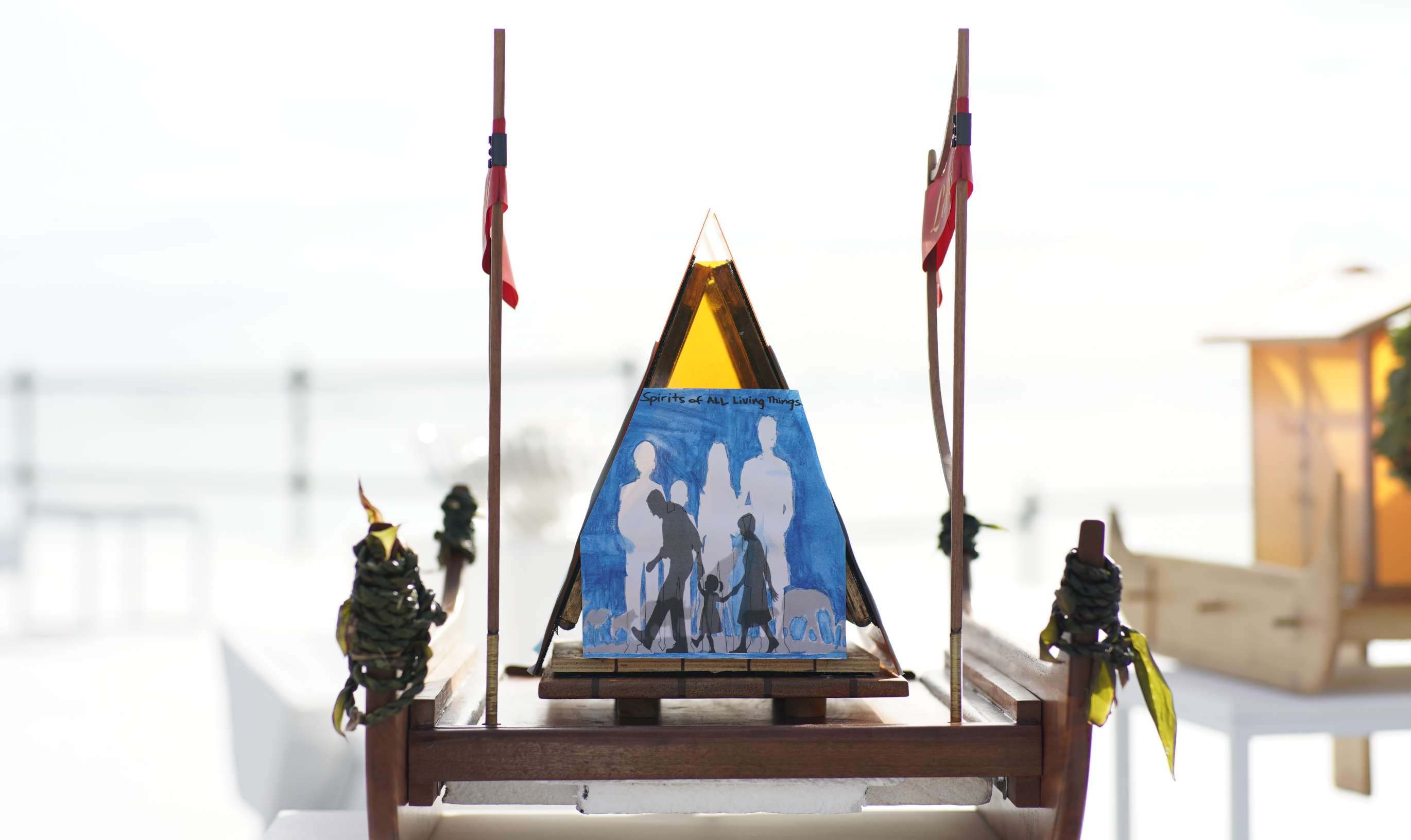 A large lantern for floating designed to look like a traditional double-hulled Hawaiian voyaging canoe sits on a table; the lantern housing forms an a frame, with outer walls covered with hand-painted artwork that depicts silhouettes of young and old walking hand in hand.