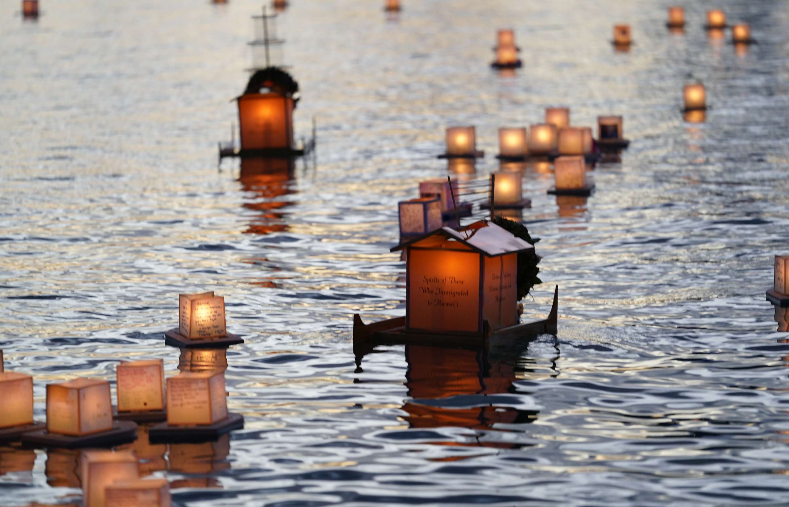 Two large, double-hulled floating lanterns made of wood and paper, illuminated from within, float on the gentle waves of a twilit ocean bay amid numerous smaller illuminated floating lanterns.