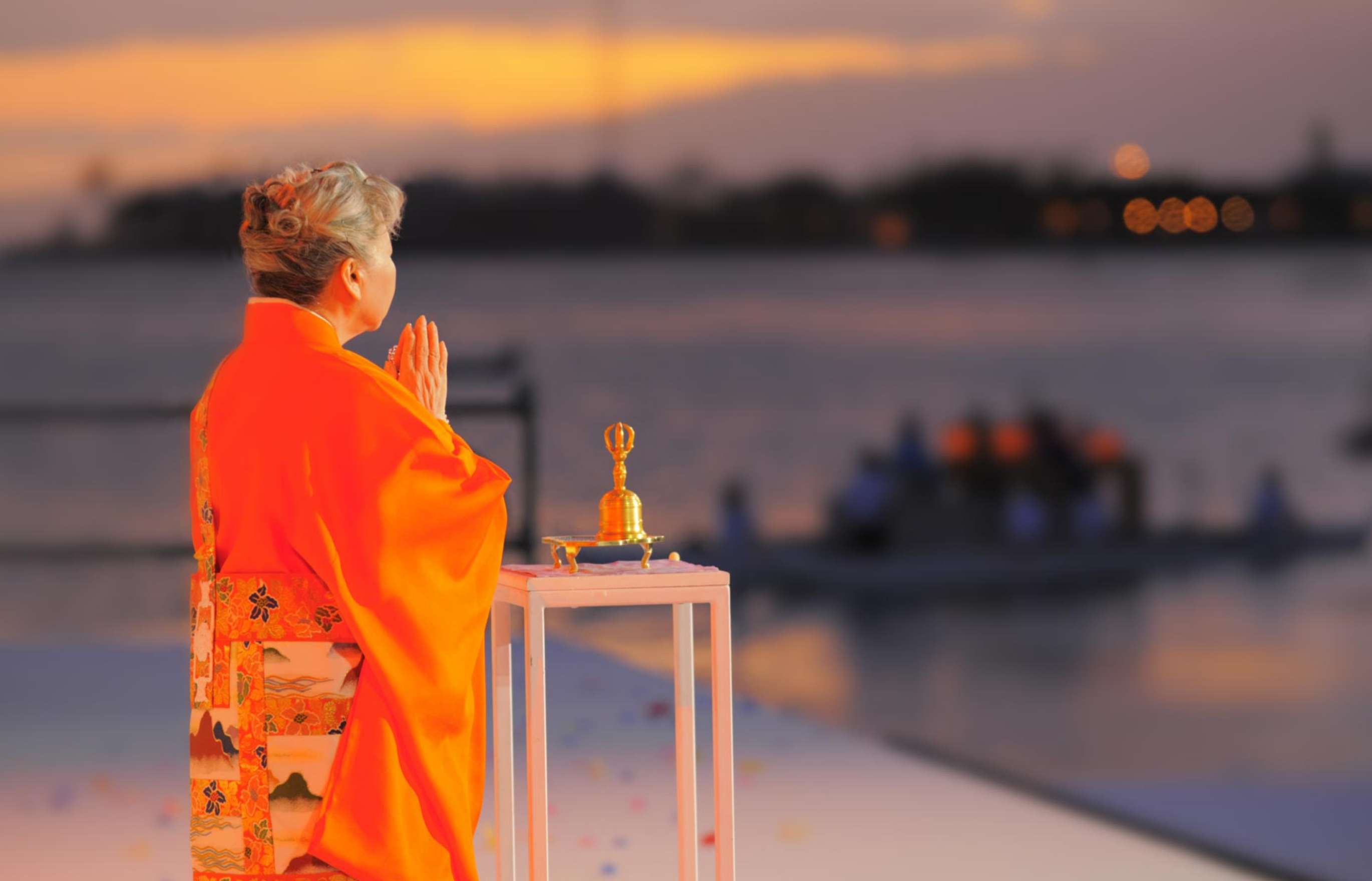 Her Holiness Shinso Ito, in bright orange robes, stands with her back to the camera before a plinth holding a ceremonial bell, her hands in gassho as she looks out over an ocean bay at sunset.