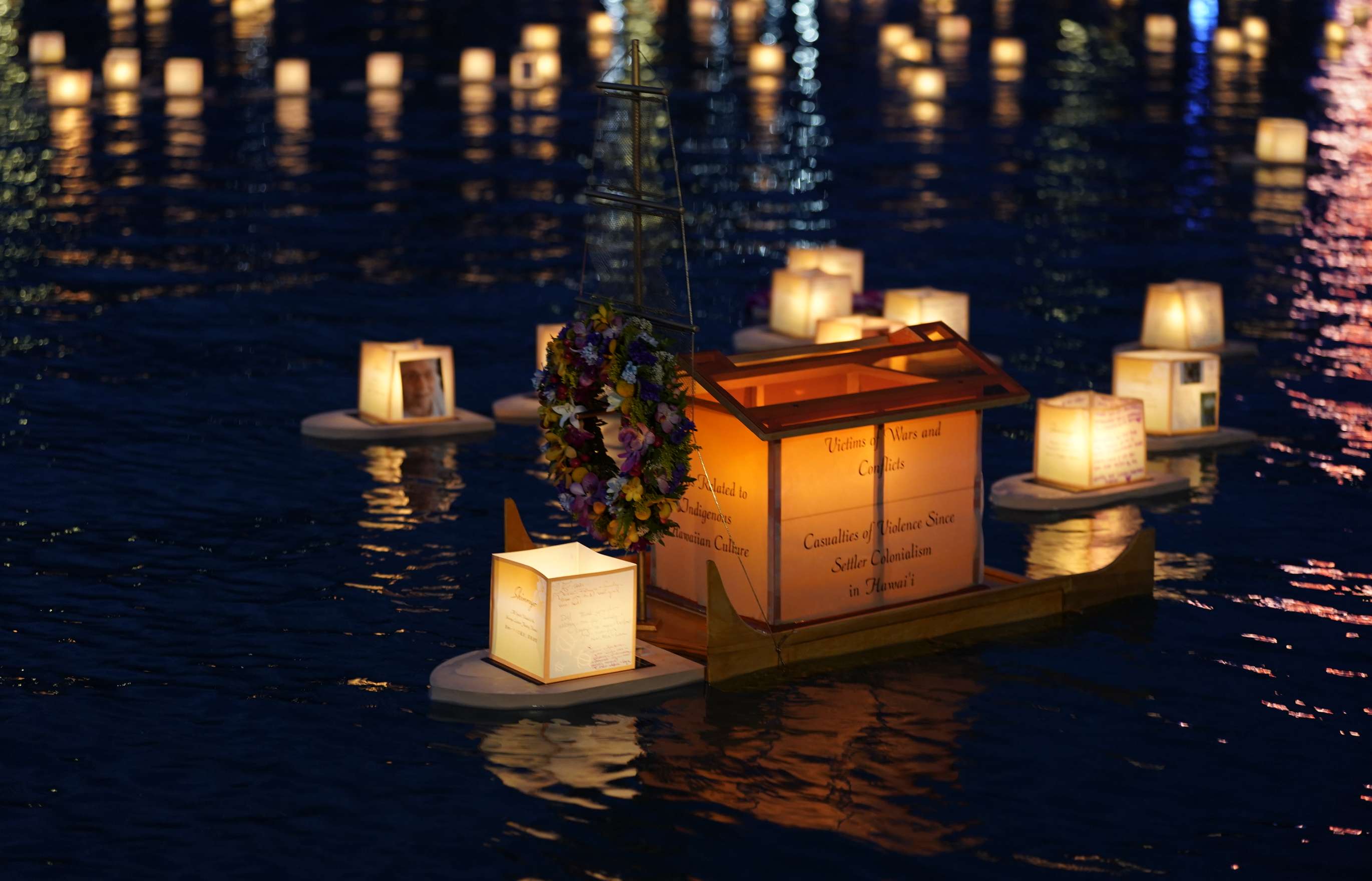 A large illuminated double-hulled floating lantern made of wood and paper, with a wreath hanging from its bow, floats amid numerous smaller illuminated floating lanterns on an ocean bay at night.