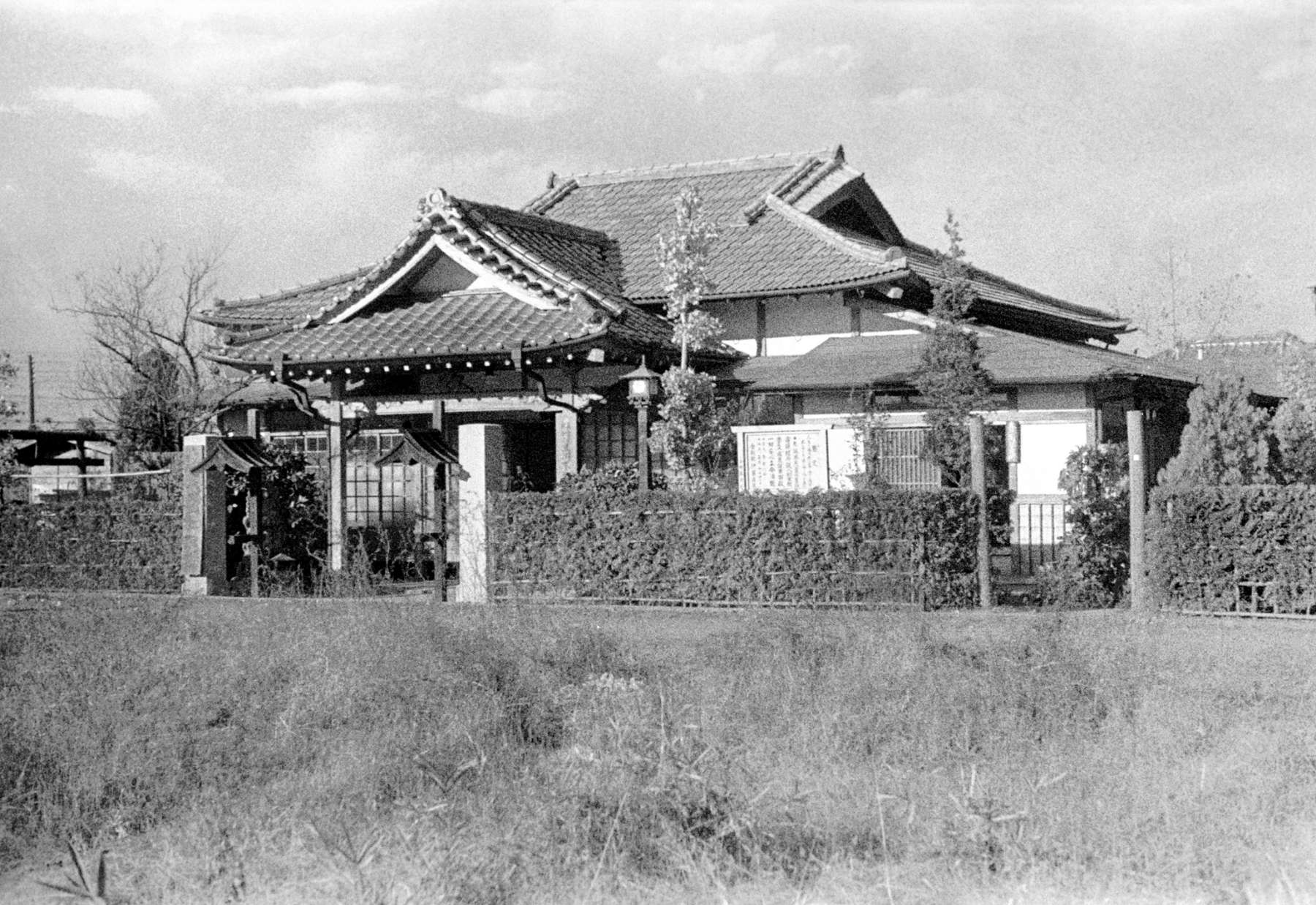A temple with tiled, peaked roofs, surrounded by hedges stands across from an empty field of grass.