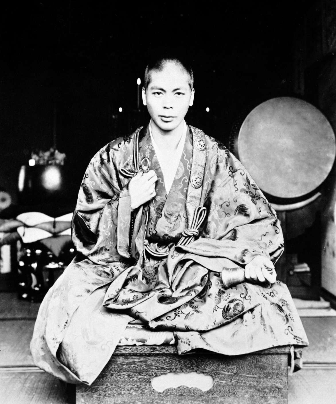 A young, shaven-headed Shinjo, facing camera, sits cross-legged, wearing brocade priestly robes, with a vajra in his right hand at his heart, and a bell in his left in his lap.