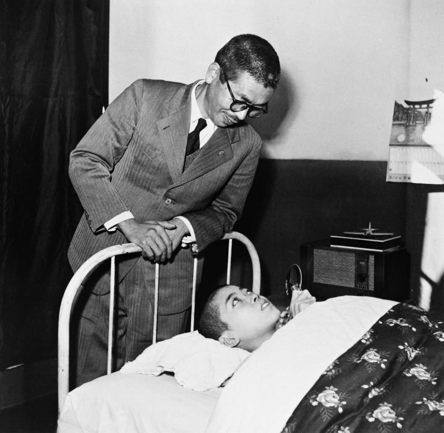 Shinjo, in a suit and glasses, leans over the head of a hospital bed, smiling at his teenage son Yuichi, who looks up from beneath a heavy comforter.