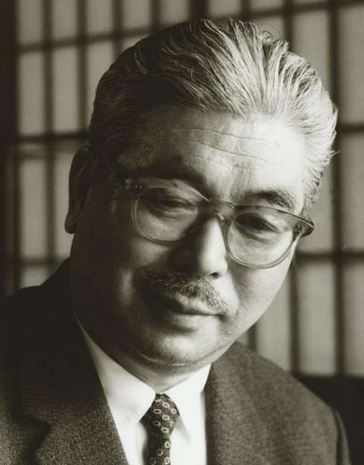 Shinjo, white haired, mustachioed, and wearing glasses, sits for a portrait in a suit, with a kindly, reflective expression on his face.