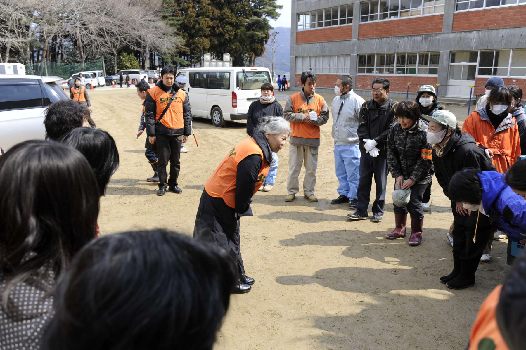Her Holiness, white haired and wearing an orange safety vest, bows to a group of Japanese people, some wearing masks and gloves, in a sand-covered parking lot.