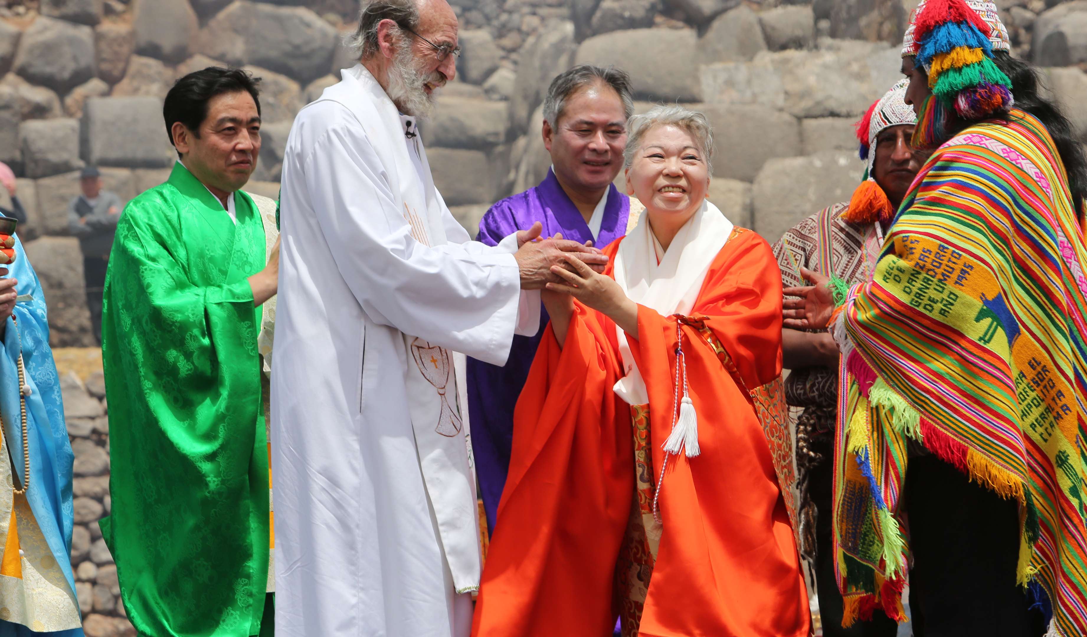 Her Holiness, in bright orange robes, clasps the hands of an elderly Christian priest in white robes as she looks, smiling, at a Peruvian man dressed in brightly colored traditional dress.