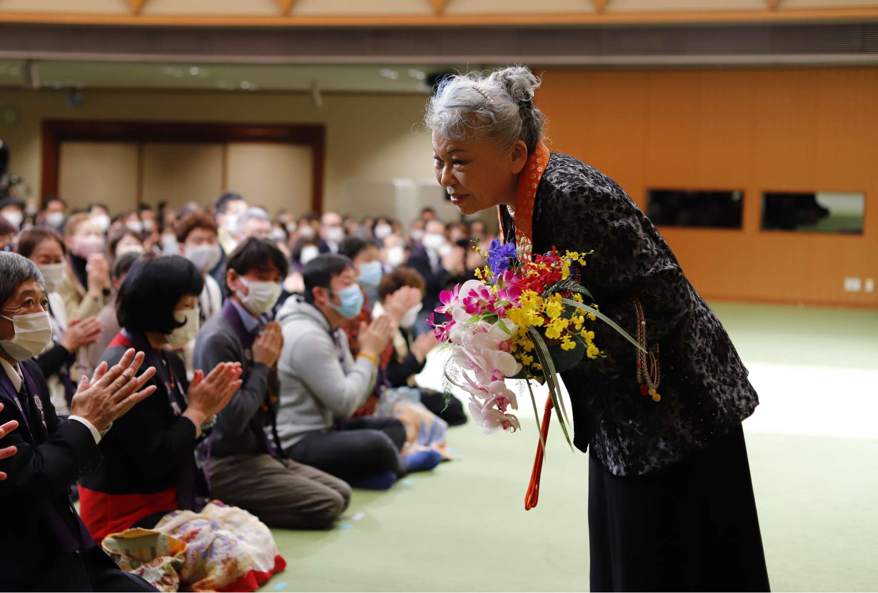 Her Holiness, in a patterned jacket and dress with bright orange kesa, holding a bouquet of flowers, bows slightly to greet seated members of a congregation.