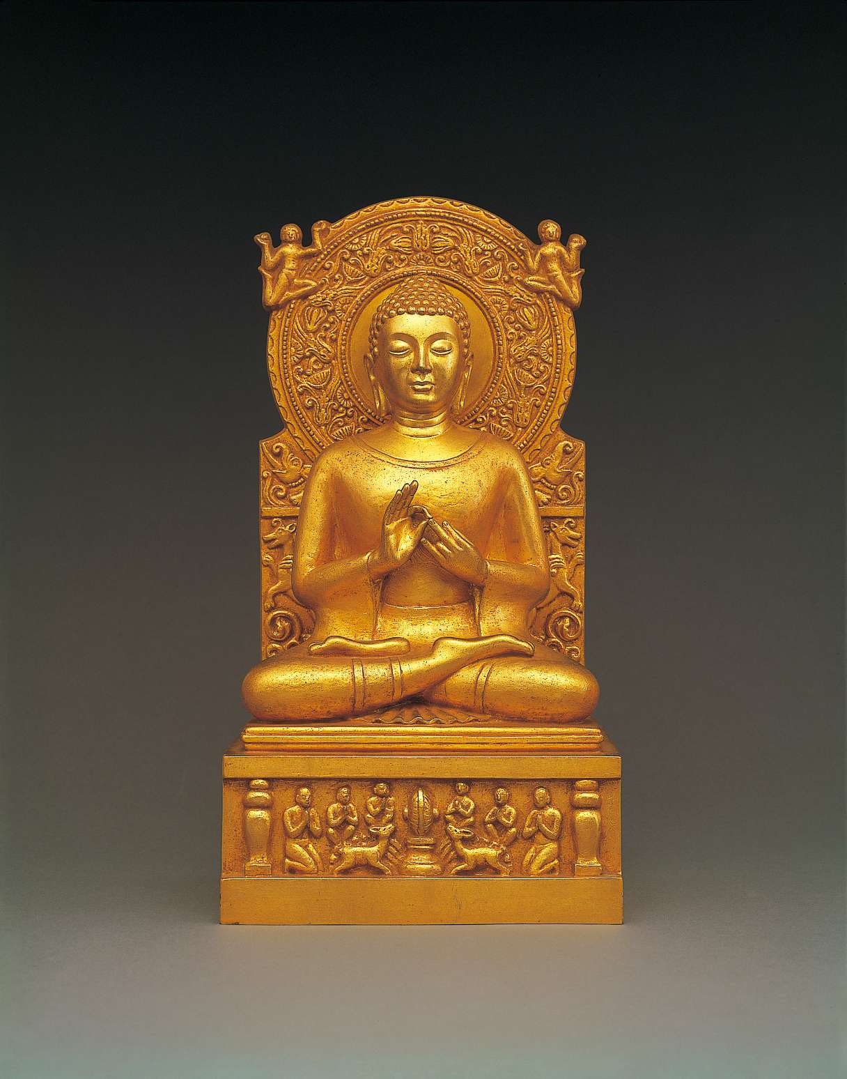 A golden hued statue of Buddha sitting cross-legged, his hands in teaching gesture, atop a throne decorated with images of disciples, deer, and a wheel, backed by an ornately decorated nimbus.