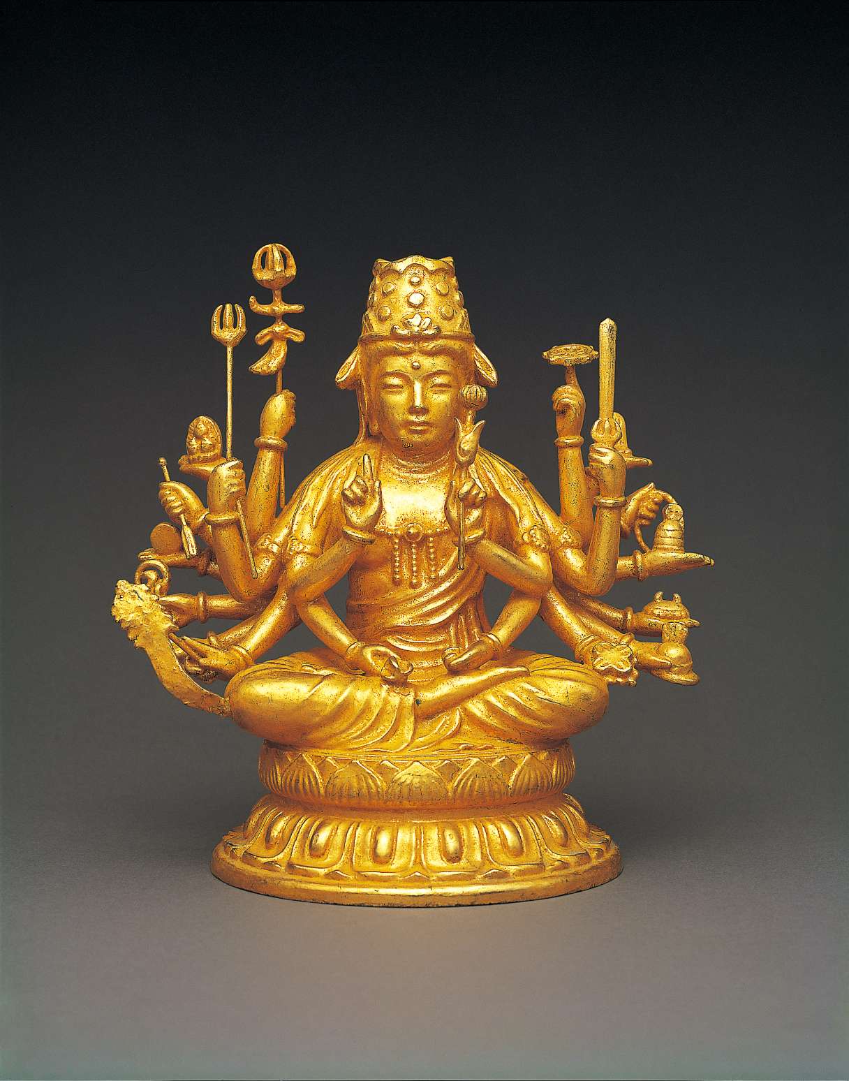 A golden hued statue of a Buddha in royal garb and jewelry, wearing a high crown, with an array of twenty arms bearing symbolic implements, sitting cross-legged atop a stylized two-tiered lotus seat.