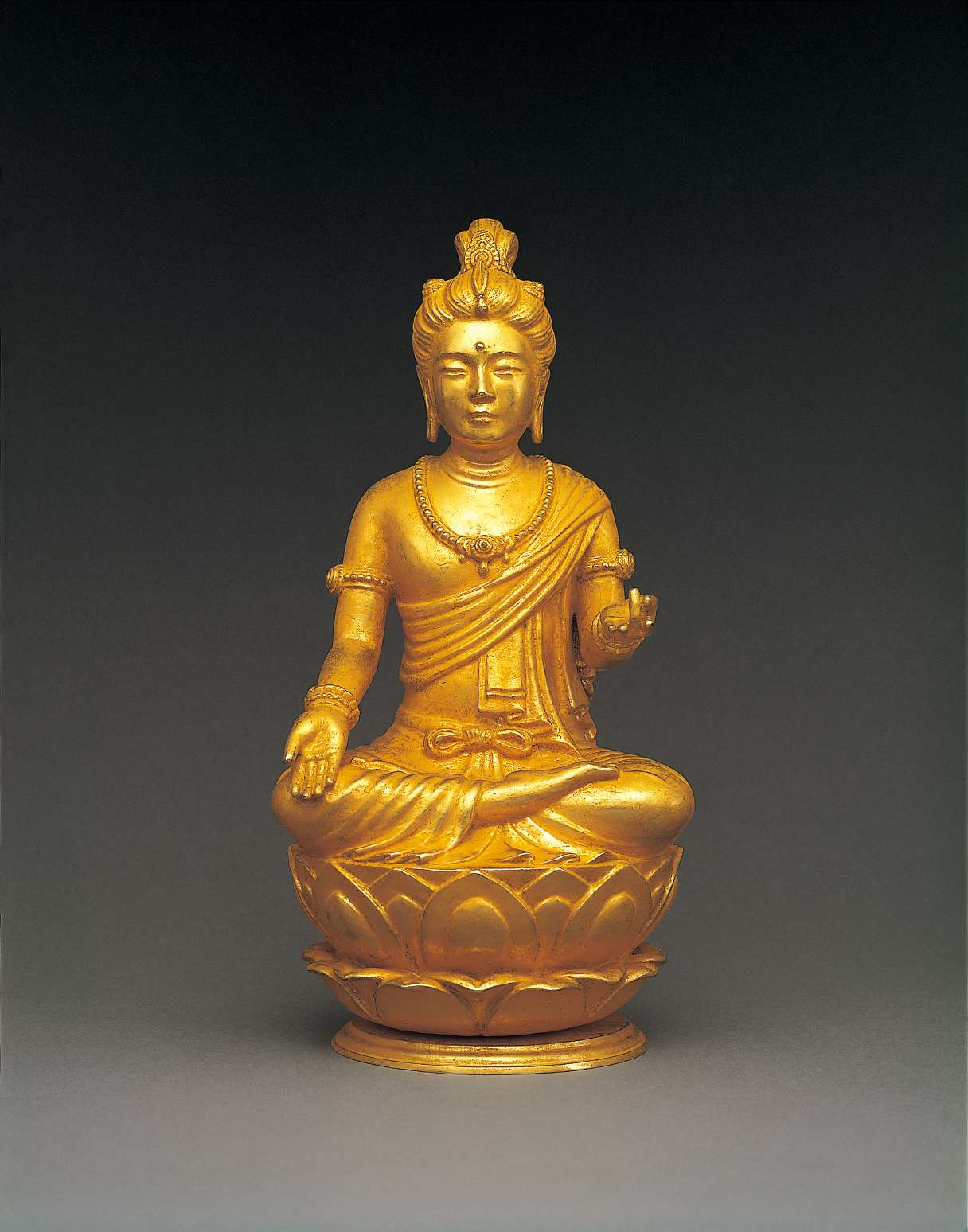A golden hued statue of a bodhisattva in royal robes and jewelry, hair in topknot, sitting cross-legged atop a lotus seat, right hand resting palm upturned on right knee, left hand raised, upturned.