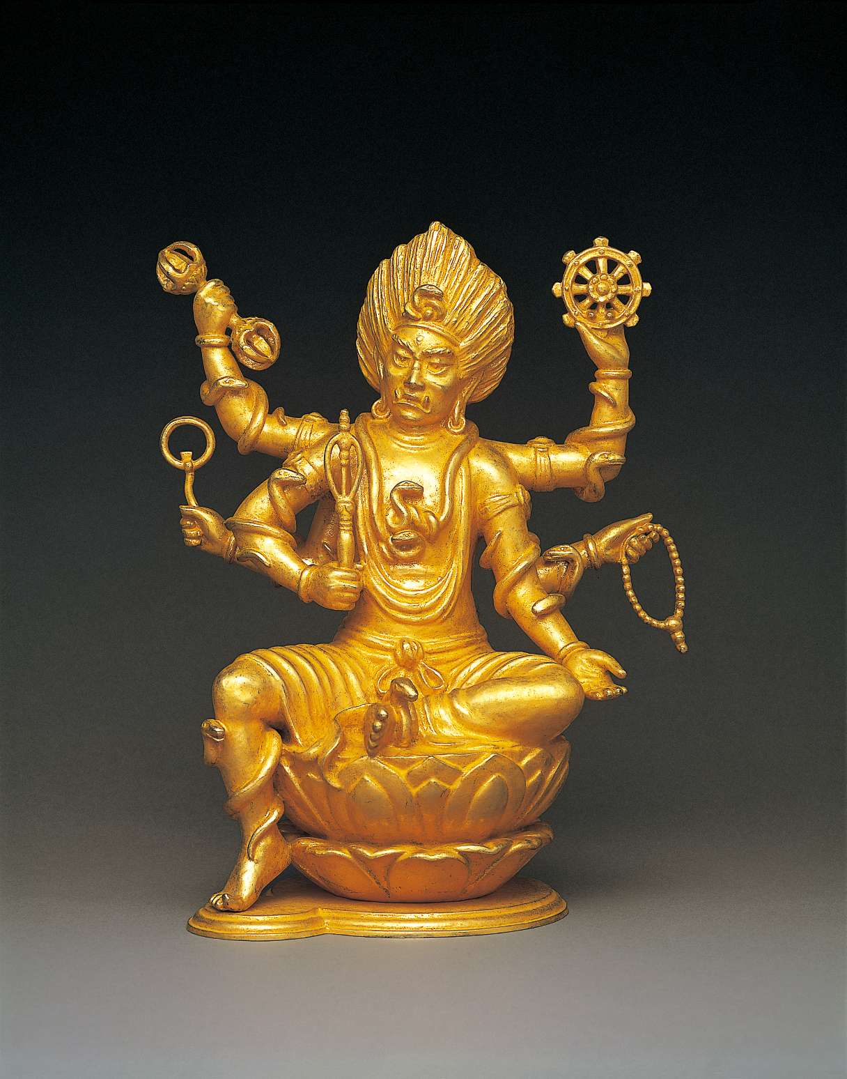 A scowling golden hued six-armed figure with fangs and wild windswept hair sits one leg akimbo atop a lotus seat; a snake coils on each limb and on his brow, and each hand wields a symbolic implement.