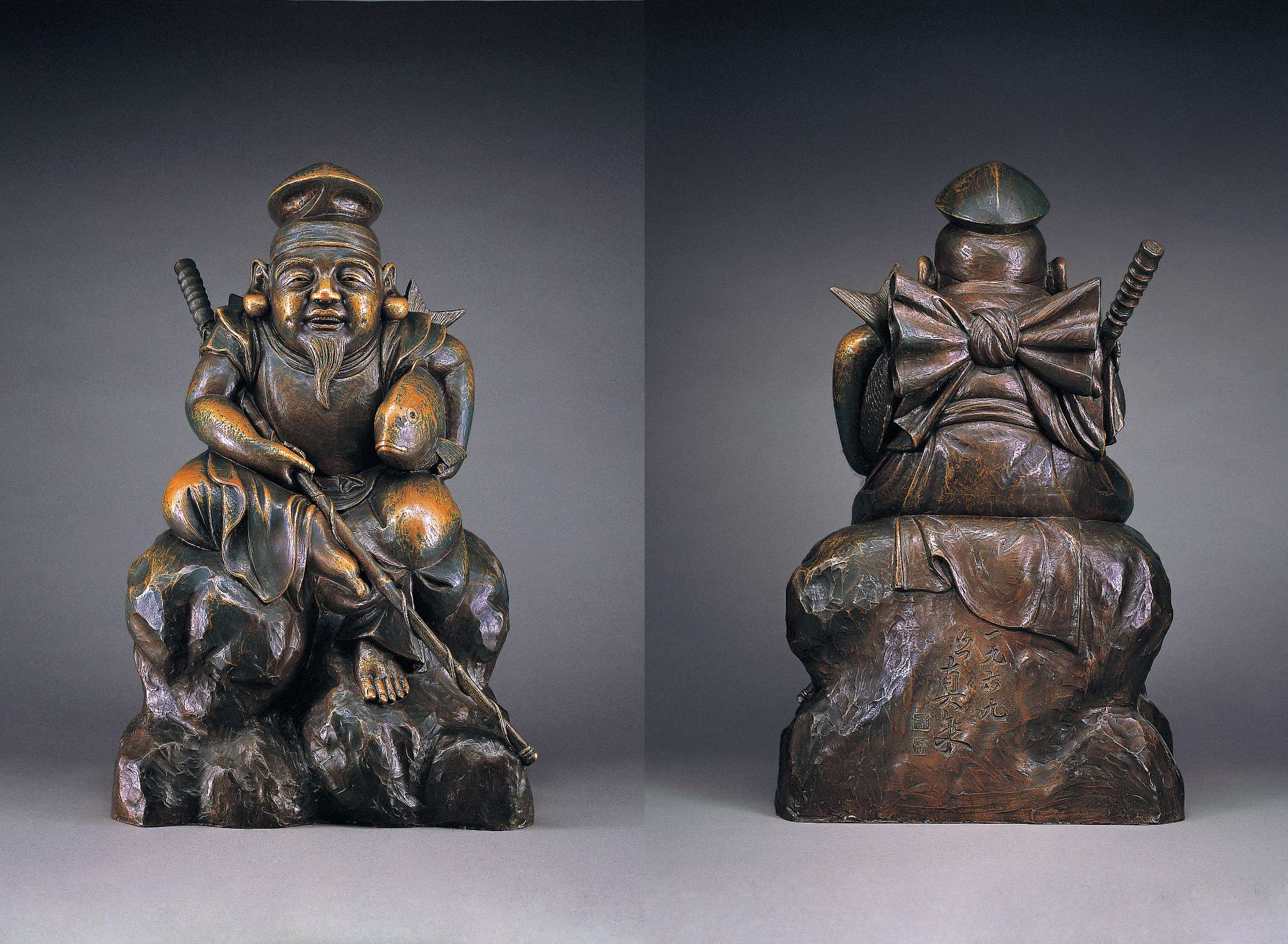 (left) A smiling, stout, bearded man in Chinese tunic and cap sits barefoot, one leg akimbo, atop a large rock, his right hand holding a long spear-like implement, his left hugging a large fish to his body. (right) The back of the statue shows the long sleeves of the man’s tunic tied in a large bow to leave his arms bare; Japanese calligraphy and stamps can be seen etched into a smooth portion of the statue.