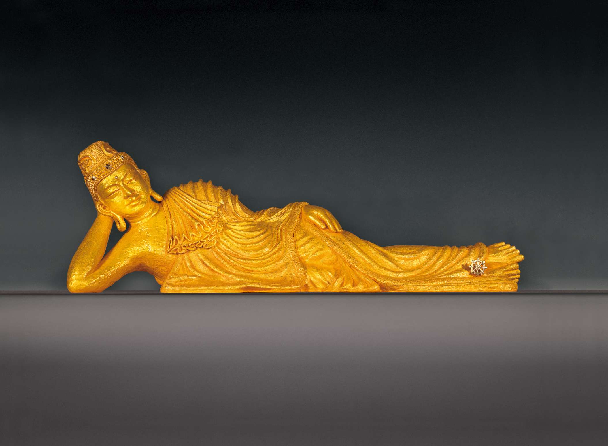 A golden colored statue of Buddha laying on his right side, head propped up by his right arm, his left arm resting on his left side, against a gradient gray background.