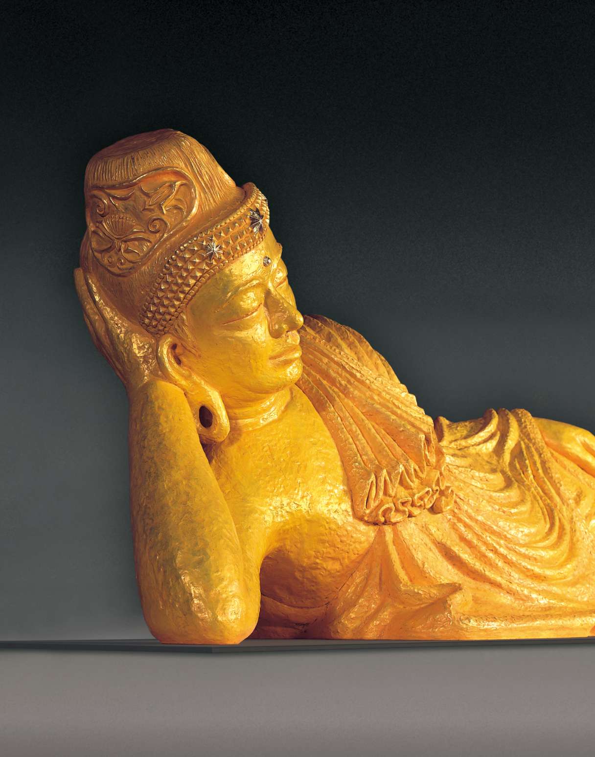 A closeup of the face of a golden colored reclining Buddha sculpture shows a slight smile on his lips; decorative details of the headpiece he wears are visible.