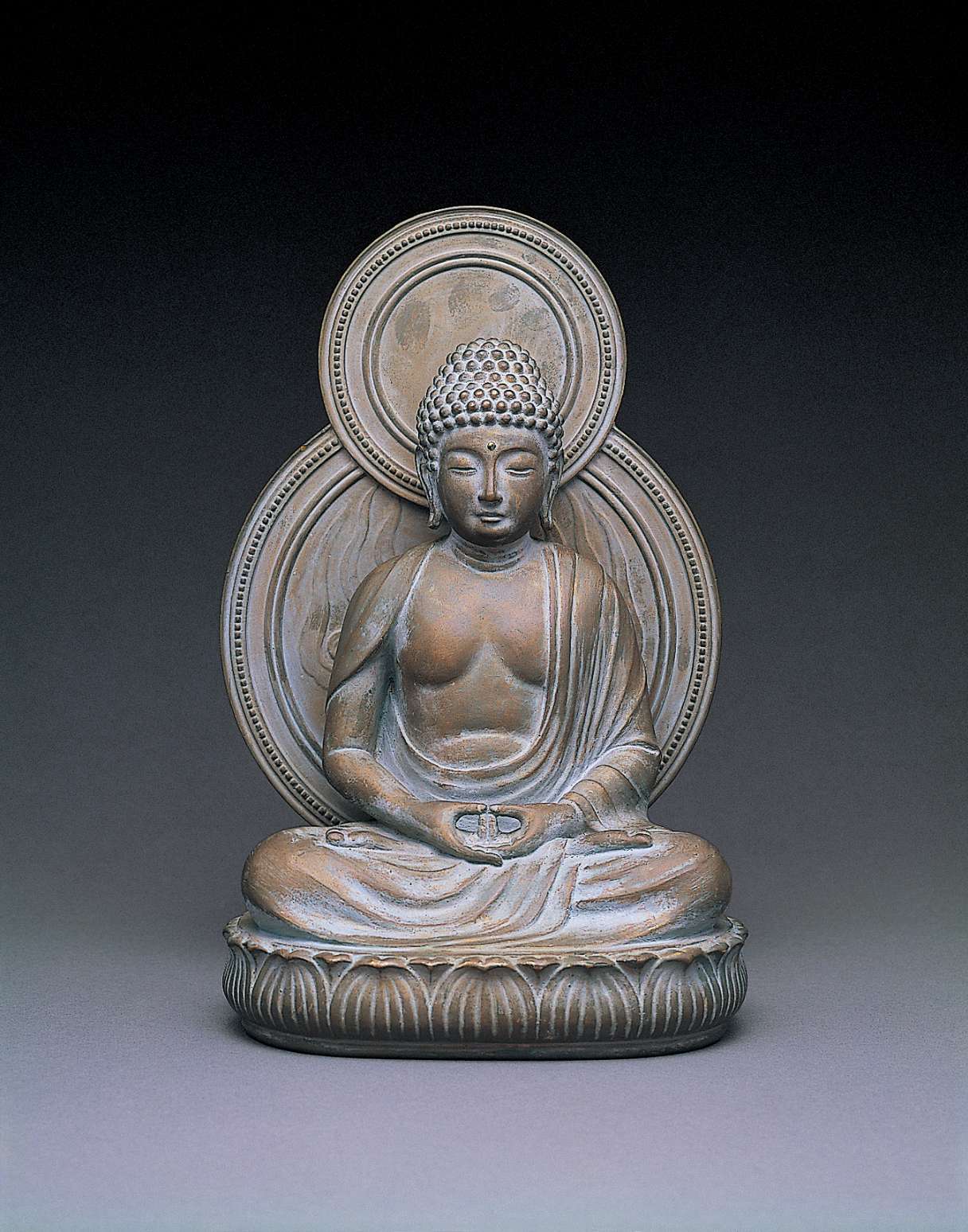 A matte, gray hued statue of a buddha seated cross-legged atop curving lotus petals, with a circular nimbus behind his body, hands in meditation. The nimbus behind the head shows some discoloration.