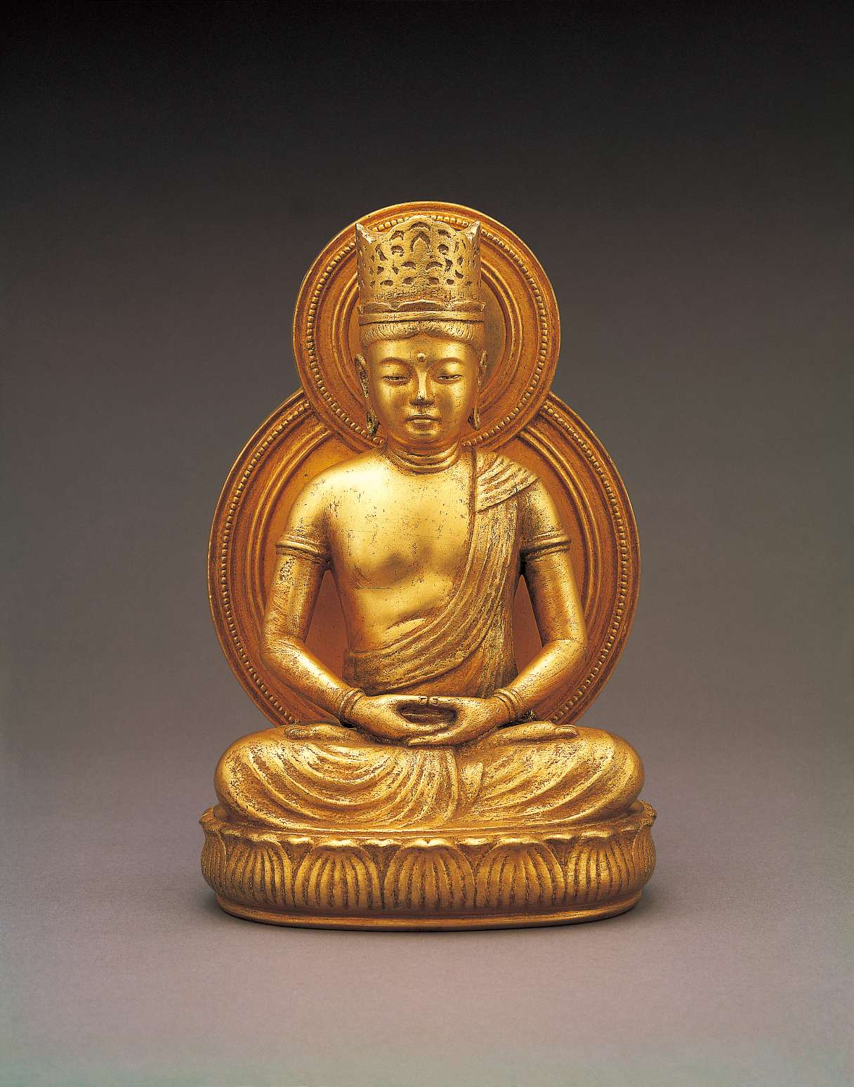 A lustrous golden hued statue of a buddha wearing a tall crown, sitting cross-legged atop a seat of curving lotus petals, hands in meditation, with a copper hued nimbus behind his body and head.