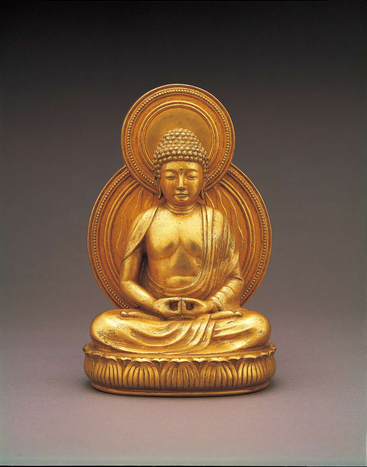 A lustrous golden hued statue of a buddha, sitting cross-legged atop a seat of curving lotus petals, hands in meditation, with a copper hued nimbus behind his body and head.