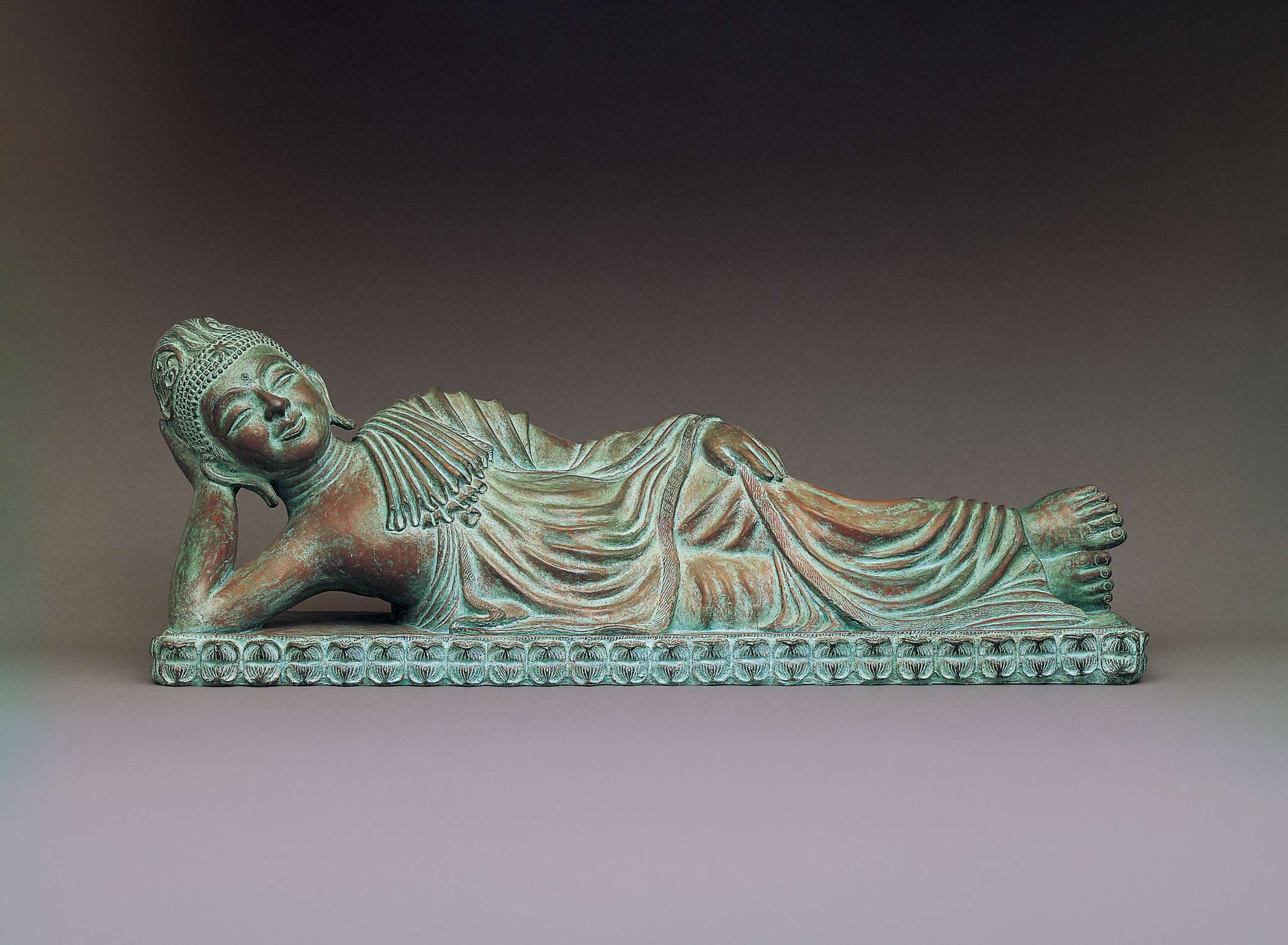 A matte metallic statue of smiling Buddha laying on his side atop a bed of decorative lotus petals, head propped up by his right arm; the drapery of his robes create a pleasing, wavelike pattern.