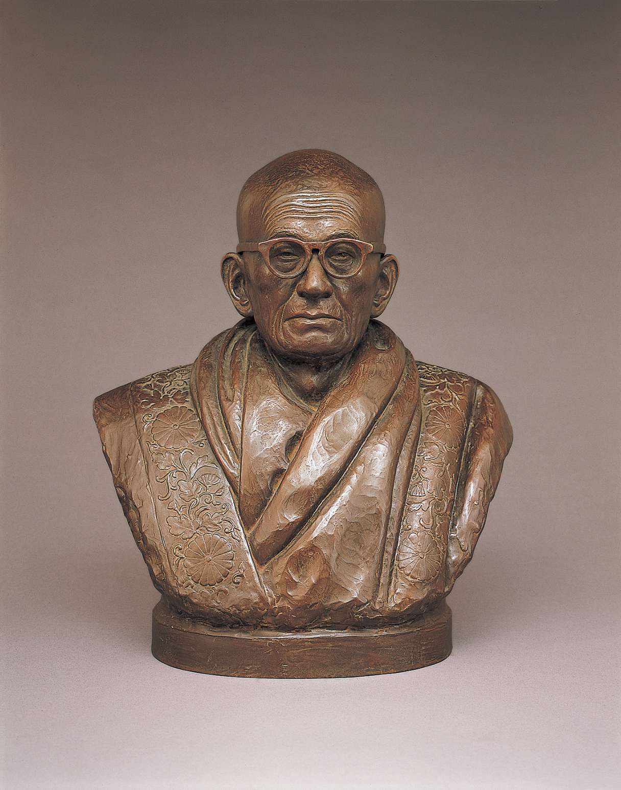 A brown hued bust of an elderly, bald Japanese man wearing horn-rimmed glasses with round frames; his face wise and lined, he wears voluminous robes over his shoulders, etched with floral designs.