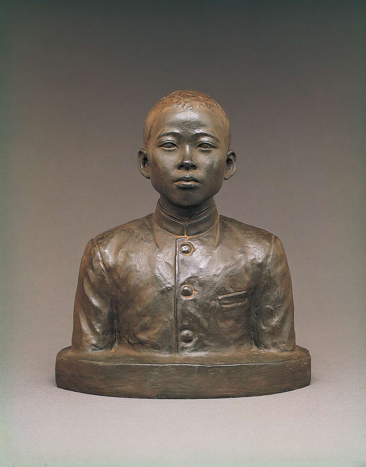 A dark gray bust of a handsome, teenaged Japanese boy with close-cropped hair wearing a buttoned up school uniform jacket. His gaze, straight ahead with eyes opened, gives a look of anticipation.