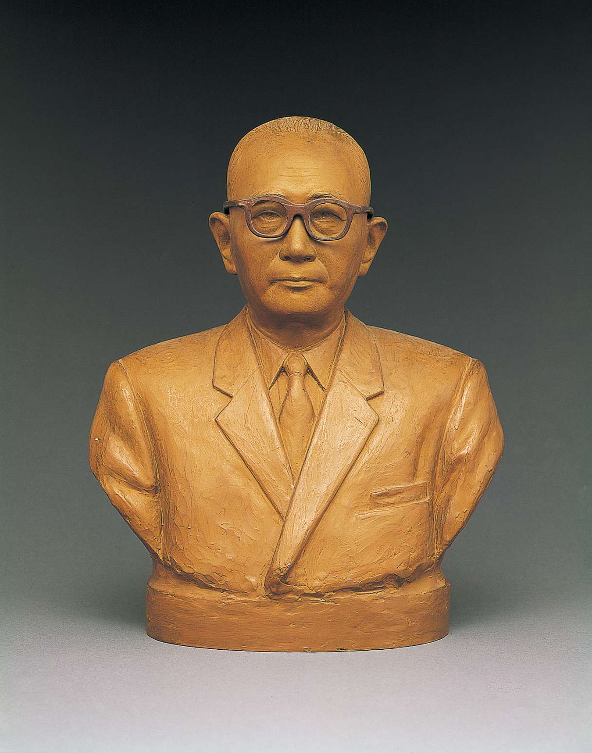 A beige hued bust of a spectacled, middle-aged Japanese man wearing a suit and tie, his face shows a kindly gaze with a determined expression.