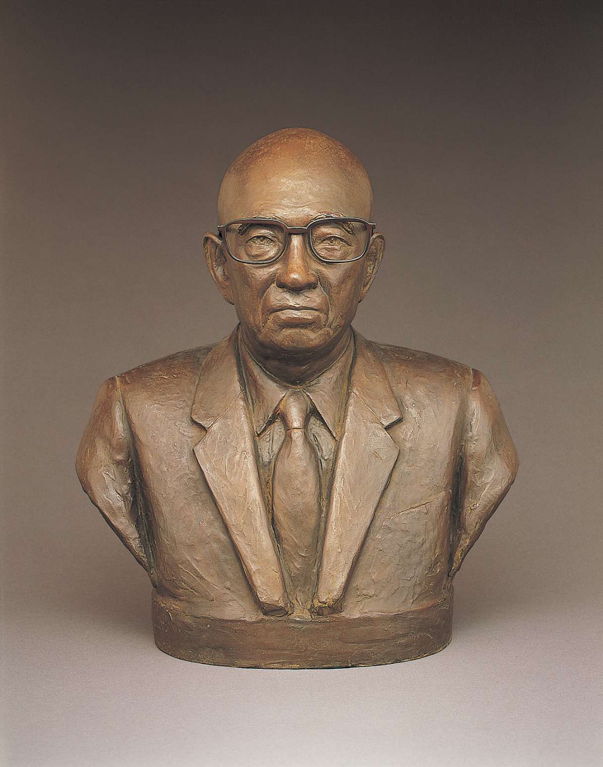 A brown hued bust of an older, bald Japanese man wearing glasses with large rectangular frames, dressed in a suit and tie. His eyes gazing directly ahead impart a look of dignified determination.