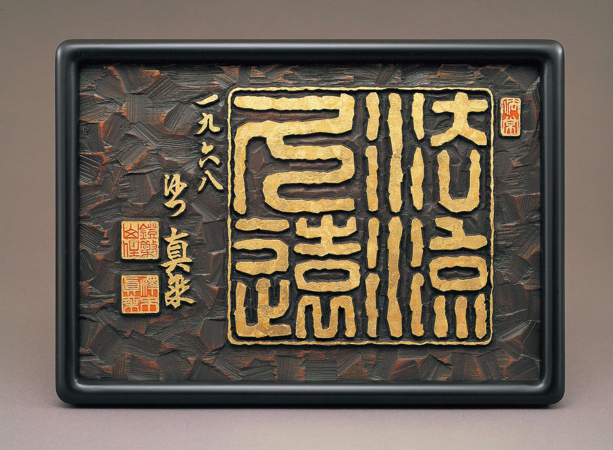 Stylized, thick-lined Japanese characters in gold are carved into a slab of dark brown wood, framed by a thin golden border, embellished by adjacent golden calligraphy and red and gold seals.