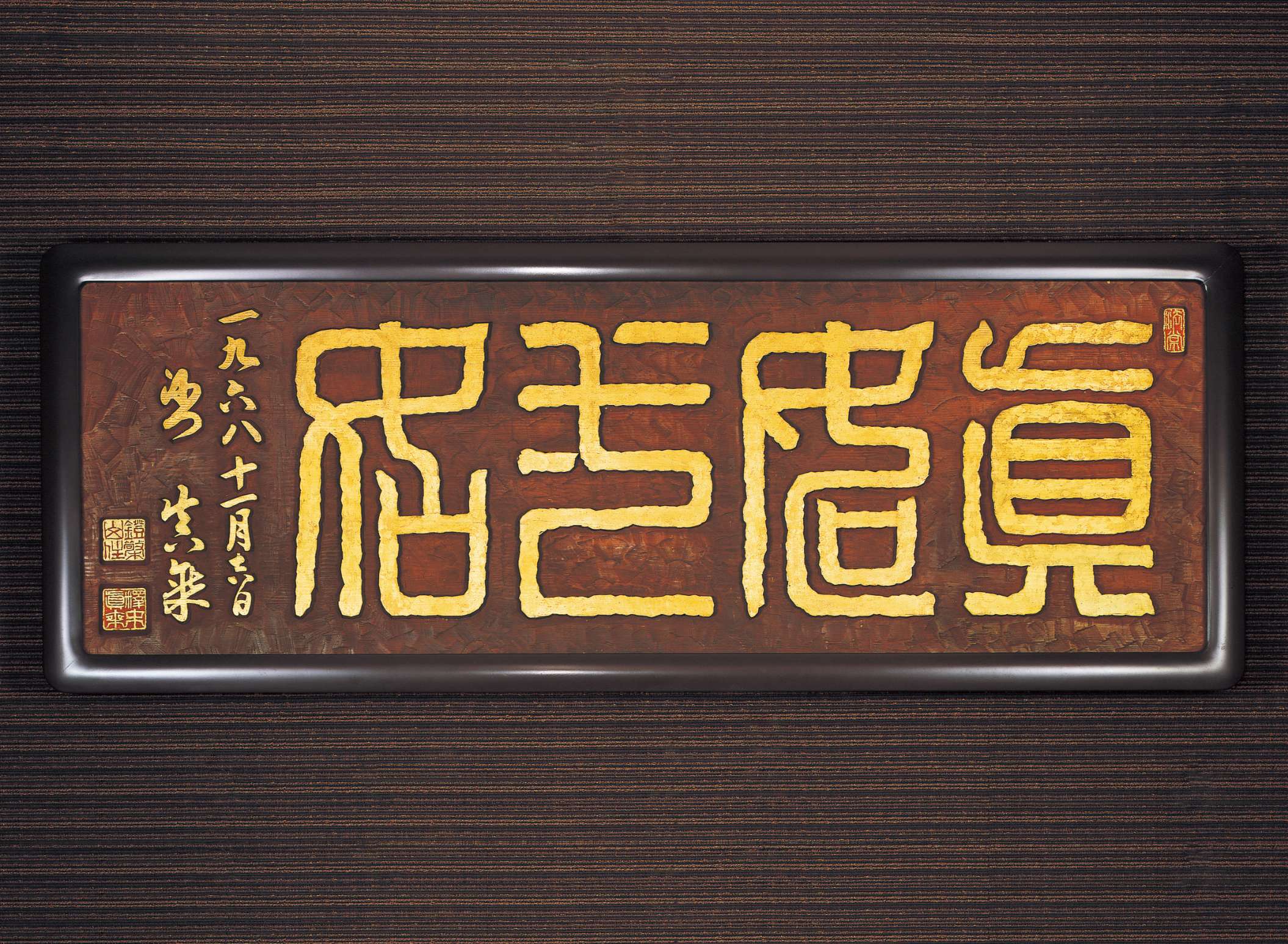A long rectangular slab of umber wood is carved with stylized thick lined, golden kanji characters that nearly fill the space, with small, vertical Japanese calligraphy in gold and red and gold seals.
