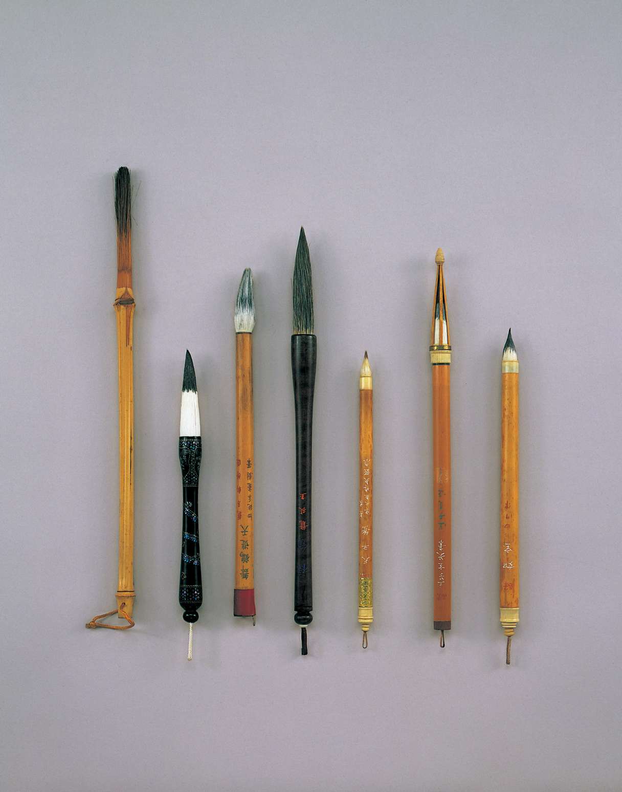 A color photograph depicts seven ink brushes of varying thickness and length, some bamboo, others of wood, marked with Japanese writing, neatly laid next to one another.