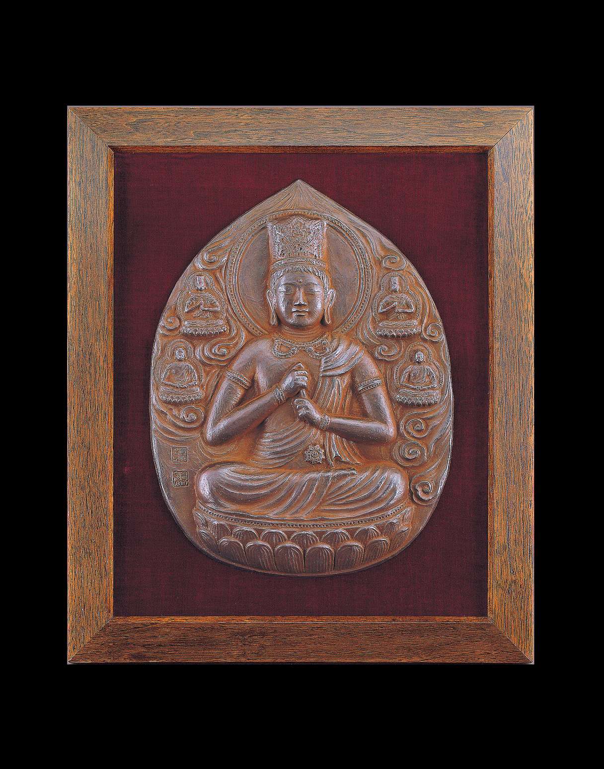 A tear-shaped metallic relief of a buddha wearing a tall crown sitting cross-legged, grasping his upturned left index finger with his right hand, surrounded by clouds and four smaller buddhas.
