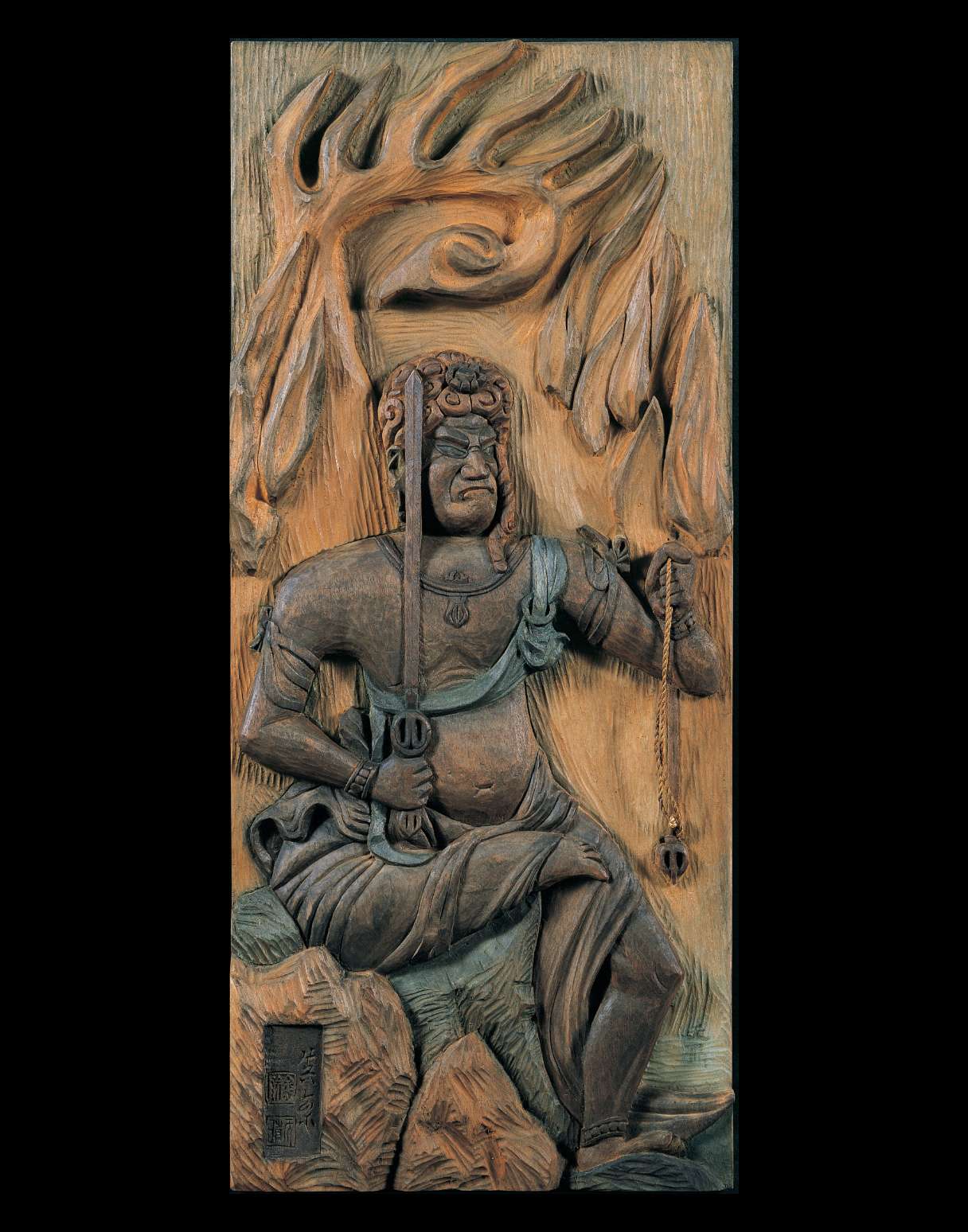 A dark relief image of a scowling figure, seated with one leg akimbo, wearing a sash, jewelry and lower robe, holding a sword in his right hand and noose in left, is carved into a reddish-ochre slab.