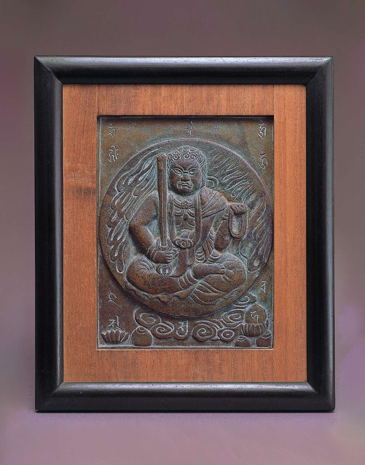 A dark, metallic relief of a scowling figure holding a sword in his right hand and a noose in the left, seated cross-legged, framed by a disc of stylized fire; letters of a mantra are etched nearby.