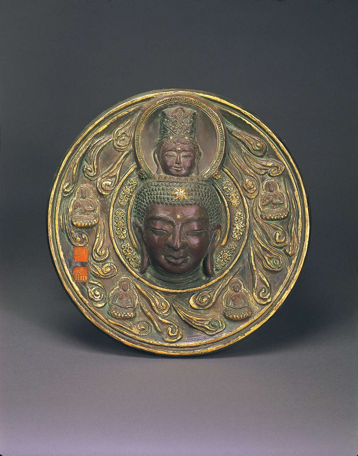 A metallic disc bears the head of a buddha at the center, atop which sits another smaller buddha head wearing a tall crown; reliefs of decorative clouds and small seated buddhas surround the heads.