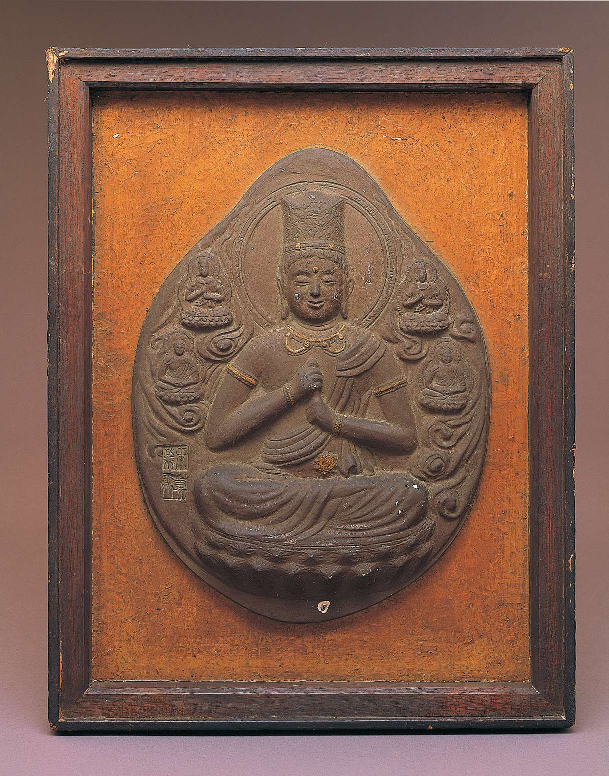 An upright almond-shaped brown relief of a buddha wearing a tall crown sitting cross-legged, grasping his upturned left index finger with his right hand, surrounded by clouds and four smaller buddhas.